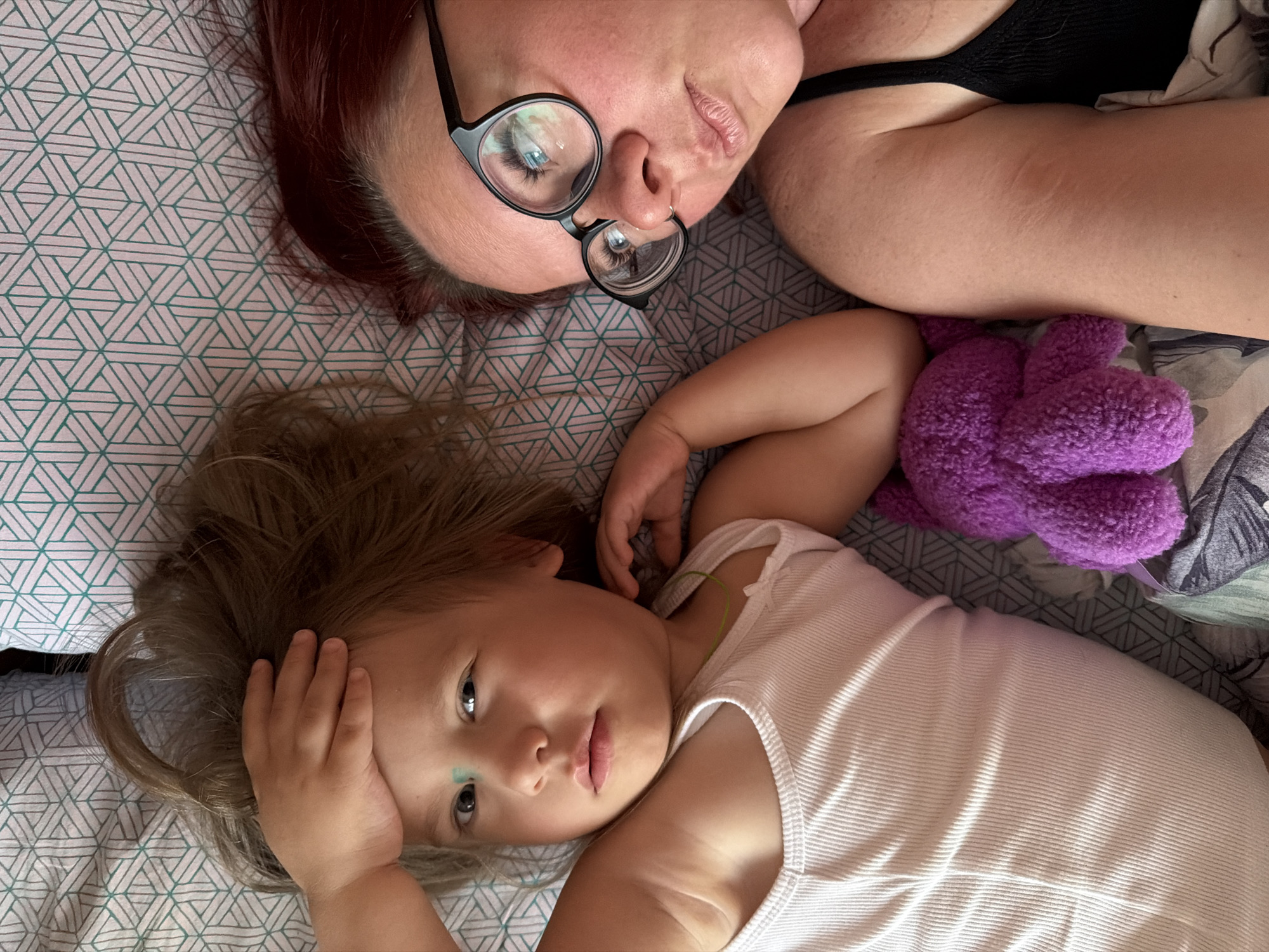 A woman and a child are lying on a bed. The woman has red hair, wears glasses, and a black top. The child has blonde hair and wears a white top. A purple stuffed animal is next to the child. The bedding has a geometric pattern in teal and white. The location is Chaikovsky, Russia.