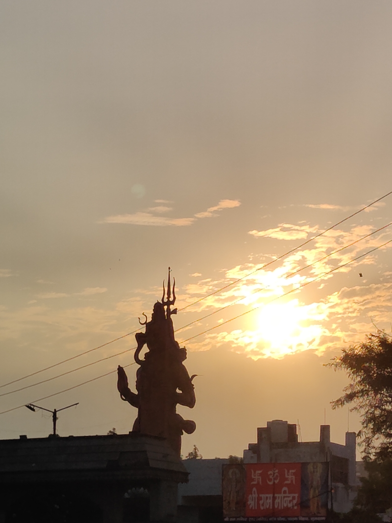 The image captures a sunset in Gurgaon, India, with a prominent statue of Lord Shiva silhouetted against the sky. The sun is a bright, glowing orb partially obscured by clouds, casting a warm, golden light. The statue, tall and imposing, features the trident (Trishul) and the serpent around its neck, characteristic iconography of Shiva. Several power lines crisscross the frame, adding a man-made element to the natural and religious scene. In the foreground, the roofs of buildings are visible, one with a sign displaying "Shree Ram Mandir" in Hindi, indicating a religious establishment. The overall atmosphere is serene and spiritual, with the vibrant colors of the sunset enhancing the grandeur of the Shiva statue.