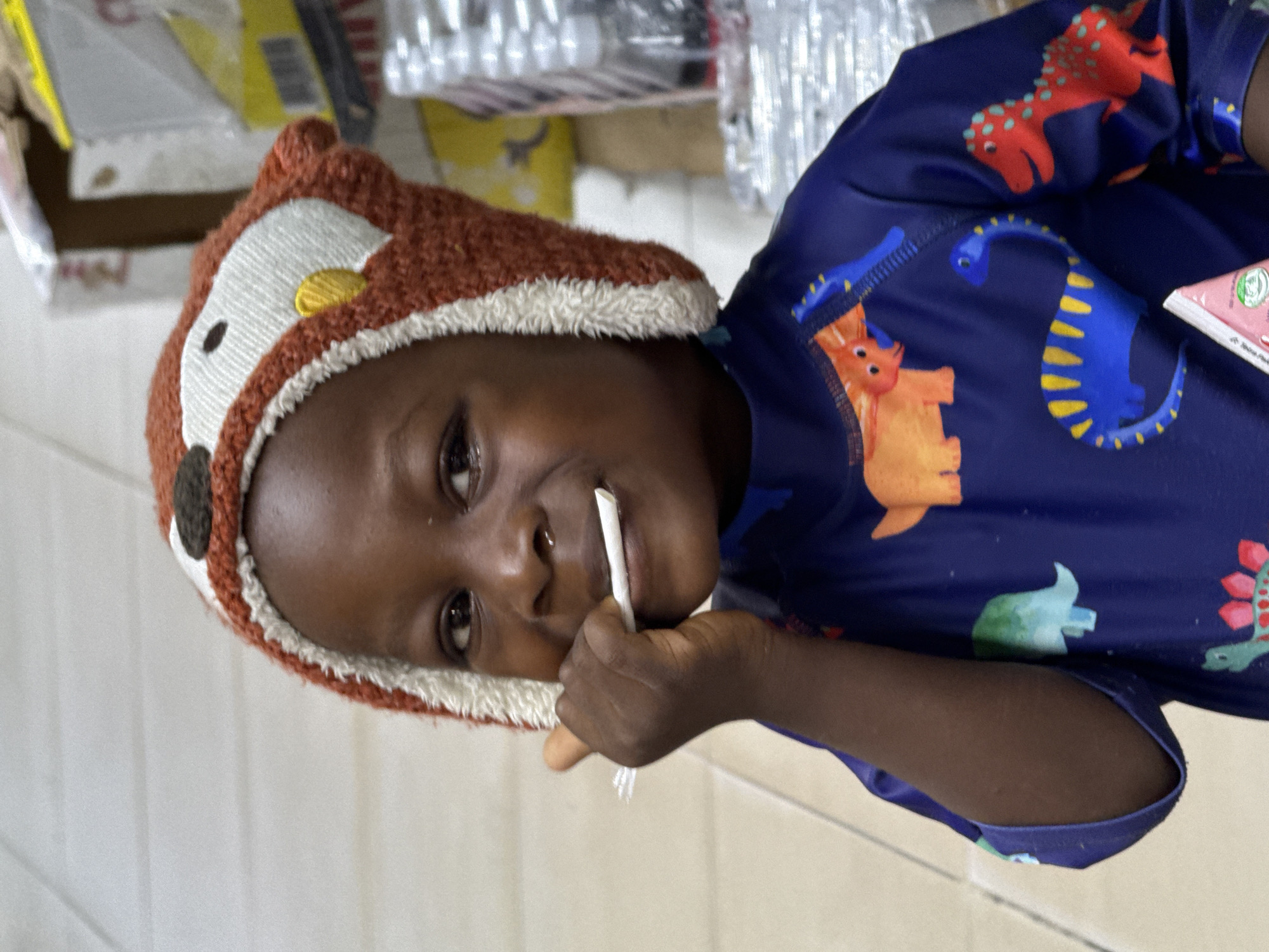 A child wearing a brown and white animal-eared hat and a blue dinosaur-patterned shirt is holding a white straw in their mouth. The child is positioned indoors, possibly in a shop, judging by the assorted packaged items visible in the background. The image was taken in Ado, Nigeria.