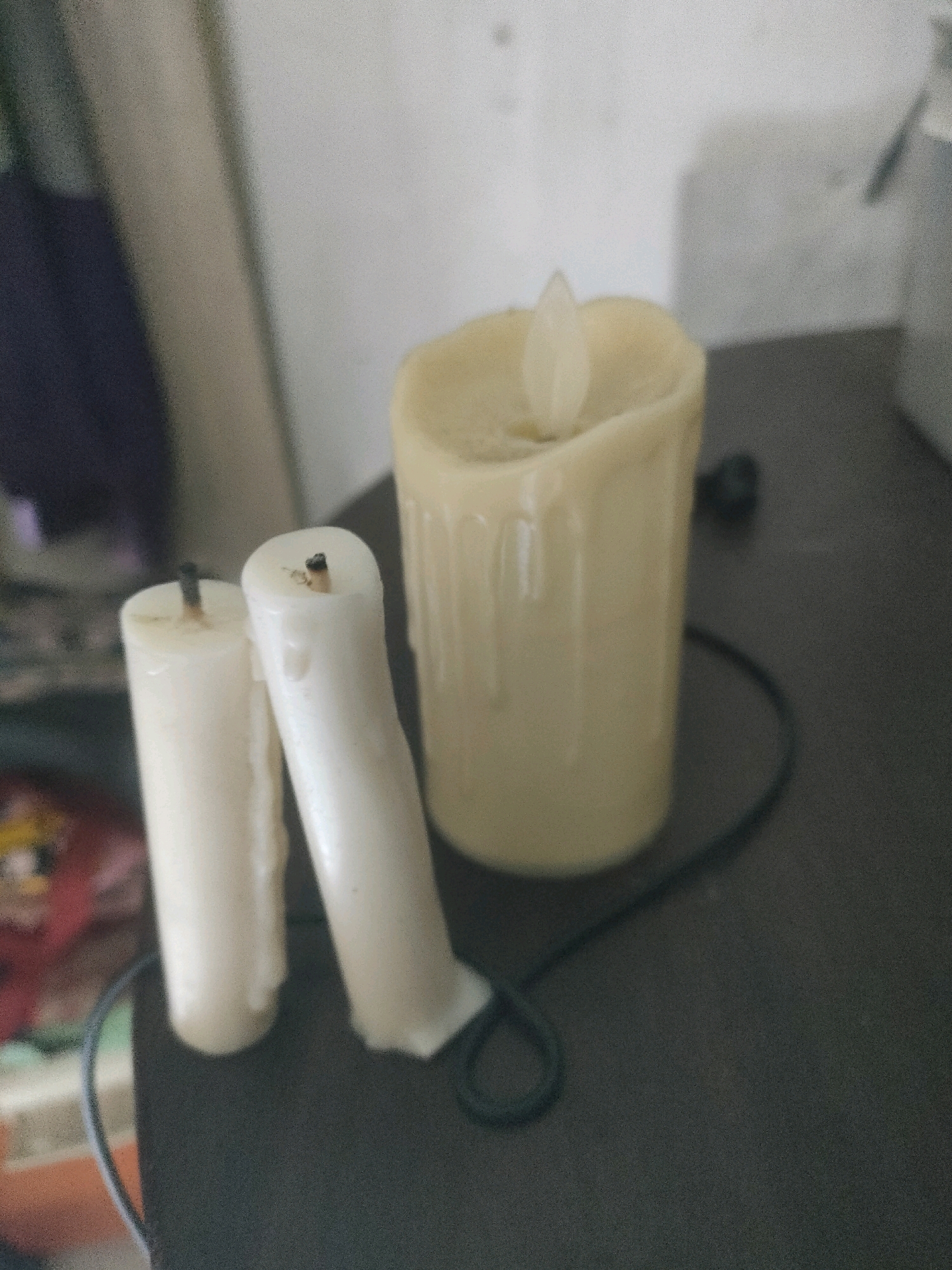 The image shows three unlit white candles, two of which are shorter and appear to be leaning against each other. The third candle is taller, thicker, and has a translucent flame-shaped piece of plastic at the top, suggesting it is an electric or battery-operated candle. The candles are arranged on a dark, flat surface, possibly a table or shelf. The background is blurred, with hints of fabric and a light-colored wall. There is a black cord snaking around the candles and disappearing into the background. The scene appears to be indoors, in a dimly lit environment. No specific location in Surabaya, Indonesia is discernible from the image.