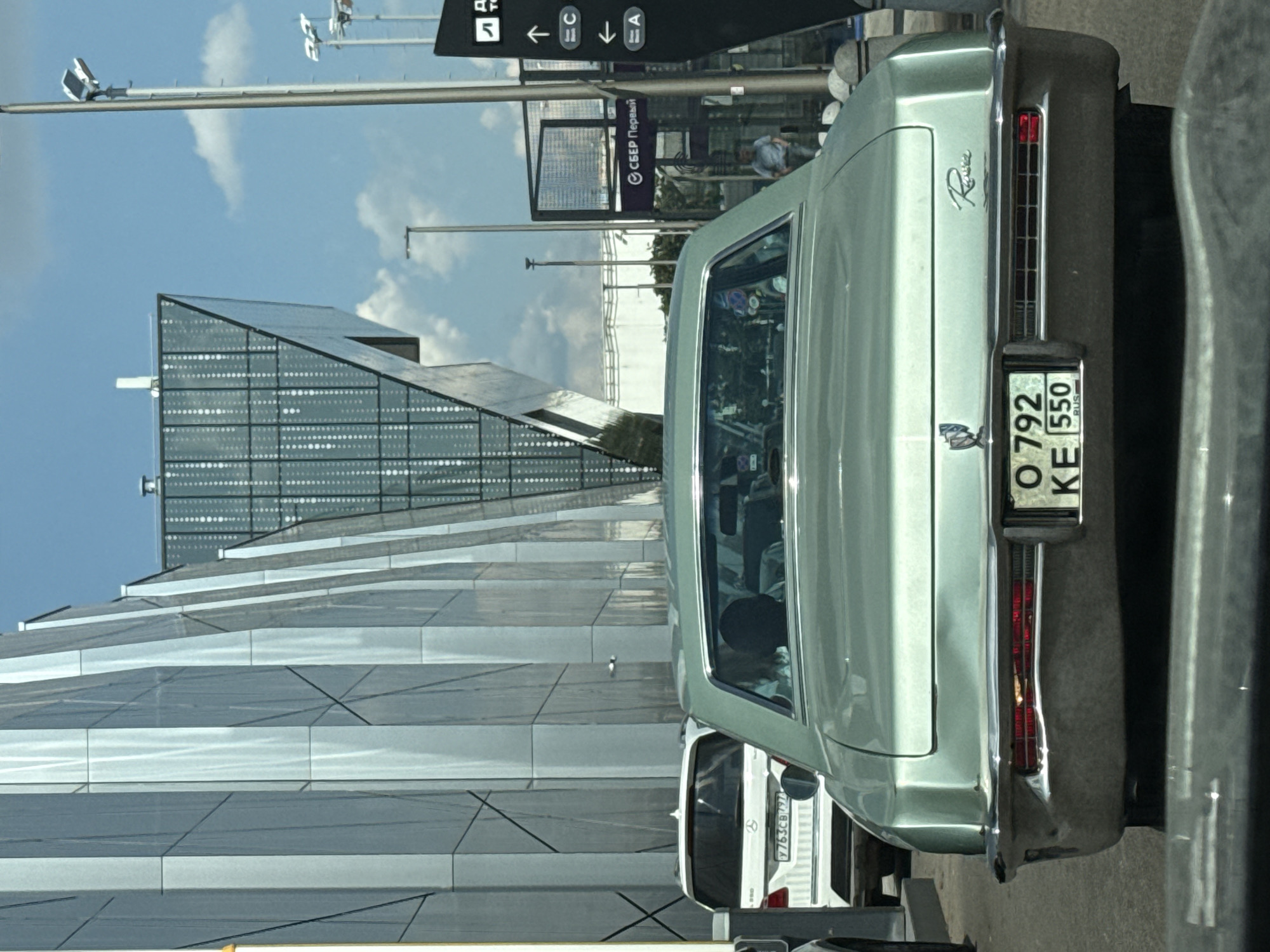 In Moscow, Russia, a pale green 1970s Buick Riviera is shown from the rear in traffic, parked beside a modern, angular building with reflective metallic panels and a distinctive triangular glass structure. The Riviera has a chrome bumper and a license plate reading "O 792 KE 550 RUS". A white Mercedes-Benz SUV is visible to the left of the Riviera. In the background, a man is walking near a sign with Cyrillic text, possibly indicating directions. The sky is blue with scattered clouds.