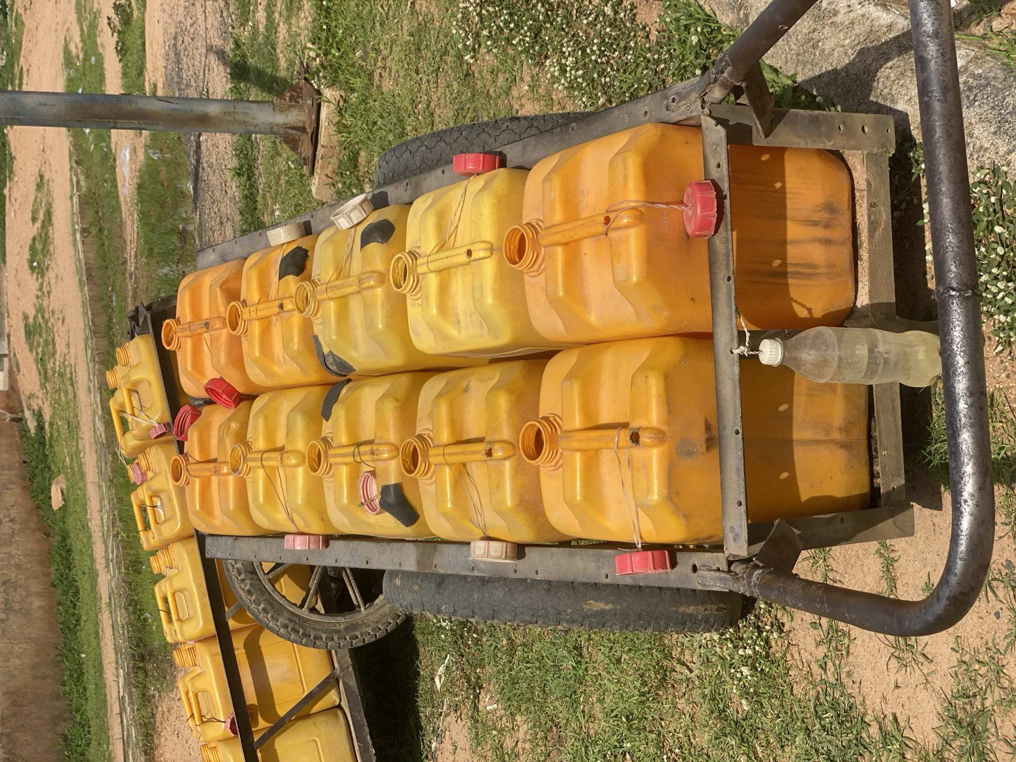 A close-up, eye-level, outdoor shot shows several rows of bright yellow plastic jerry cans, tightly packed together on a wheeled cart. The jerry cans are of various sizes and colors of caps, with some red, pink, and white. A few of the jerry cans have black markings. The cart is made of dark metal, with two large black tires. In the background, there is a dirt path, patches of green grass, and a metal pole. The scene is likely located in Jalingo, Nigeria, as indicated by the presence of the jerry cans, which are commonly used for transporting fuel in the region. The lighting suggests it is daytime.