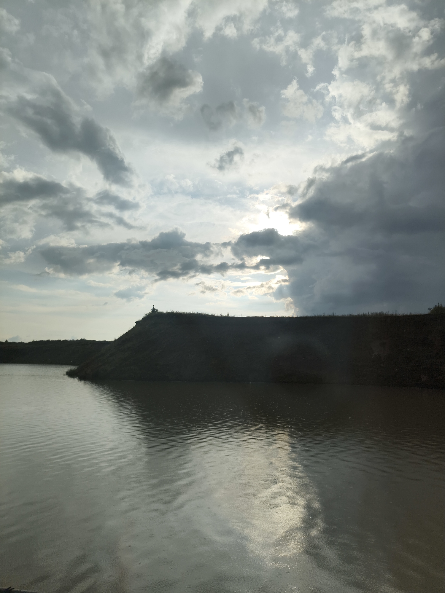 An outdoor, landscape photograph captures a body of water in the foreground and a dark, silhouetted hill against a dramatic, cloudy sky. The water is choppy, with ripples and reflections of the sky and the land. The hill, which occupies the middle ground, is covered in vegetation at its crest and slopes down towards the water. A small, solitary figure is visible standing on the crest of the hill, silhouetted against the bright sky. The sky is filled with heavy, gray clouds, with patches of sunlight breaking through in the upper right quadrant, creating a sense of depth and atmosphere. The overall lighting is dim, suggesting it may be dusk or a cloudy day.