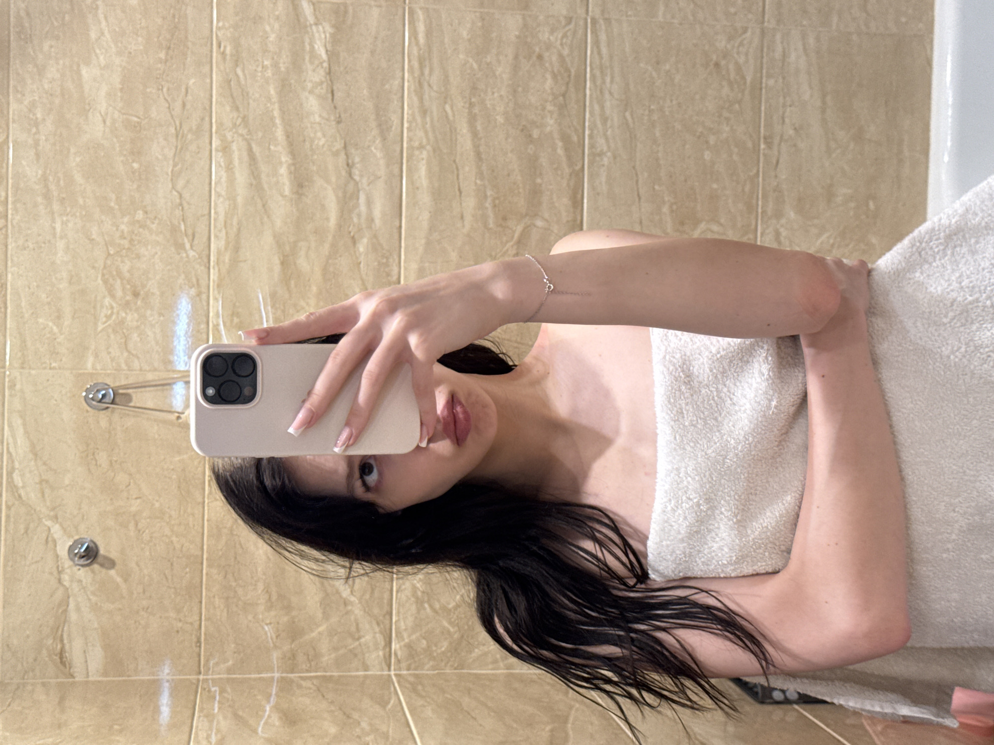 A young woman with long, dark, wet hair is taking a mirror selfie in a bathroom. She is wrapped in a white towel and is holding a phone in her right hand to take the picture. She is wearing a silver bracelet on her left wrist. The bathroom has beige tiled walls. Two chrome hooks are visible on the wall behind her. The scene appears to be a typical residential bathroom. The specific location within the city of Khimki, Russia, cannot be determined from the visual information provided in the image.