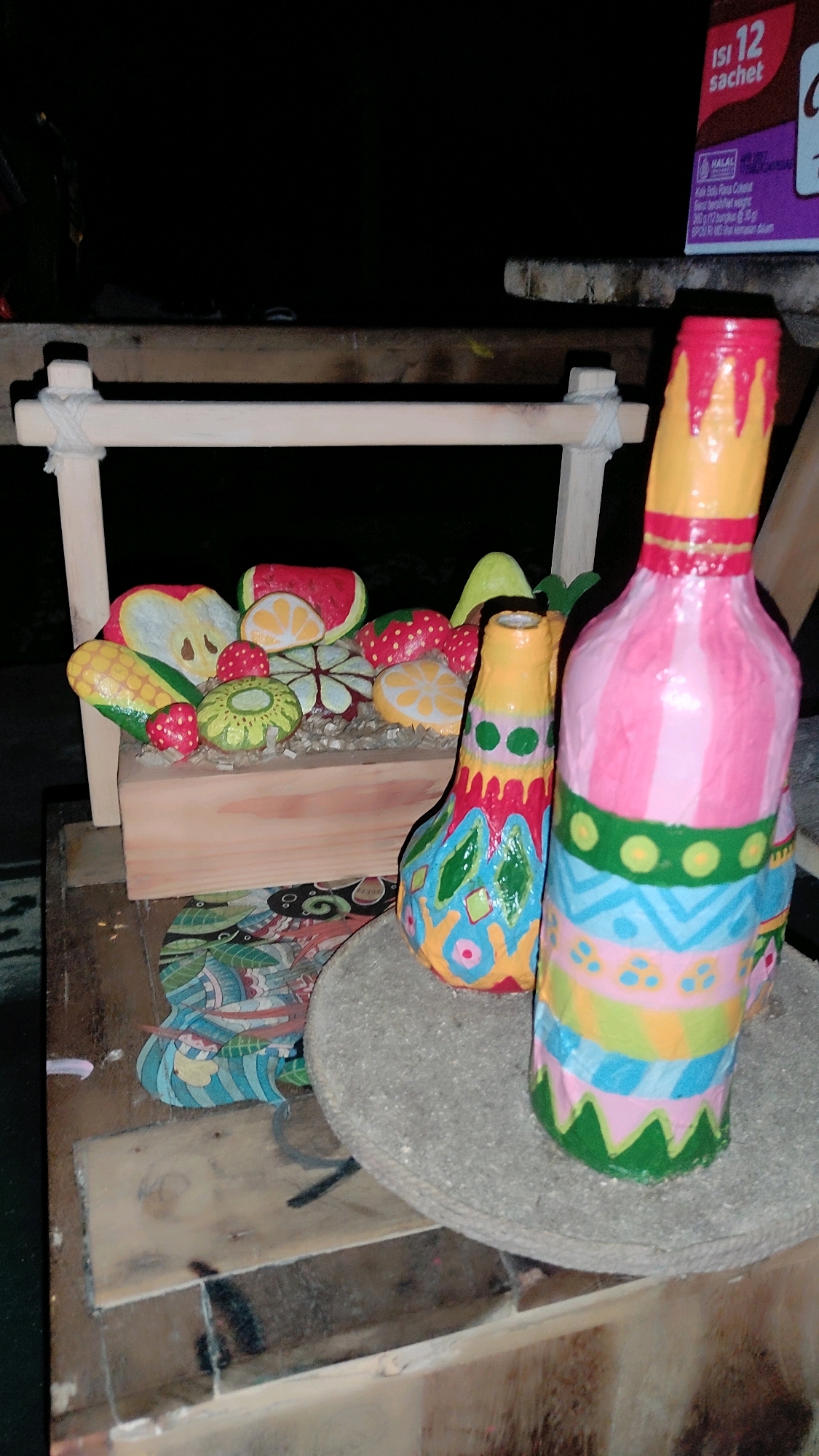 This is a medium-shot, eye-level, indoor photograph taken at night, in what appears to be an outdoor market stall in Guwosari, Indonesia. The foreground is dominated by two decorative, hand-painted bottles. The bottle on the right is taller and painted with pink, yellow, and blue stripes, along with green scalloped edges and yellow circular accents. The bottle to its left is shorter and painted with a colorful, abstract pattern of diamond shapes and zigzags in shades of blue, green, pink, and yellow. Both bottles are resting on a rough, circular, gray surface, possibly a stone base.
Behind the bottles, a small, wooden cart displays a collection of brightly painted, artificial fruits and vegetables. These include slices of watermelon and orange, strawberries, corn, and a yellow pear. The cart itself is made of light-colored wood and is supported by a wooden frame with a white rope detail. The cart is placed on a wooden table or surface.
In the background, the scene is dark, but a portion of a purple cardboard box with Indonesian text is visible on a shelf to the upper right. This box appears to be a sachet of chocolate. The overall impression is one of a lively, handmade craft display, possibly for sale at a local market. The lighting is dim, suggesting artificial light sources are being used.
