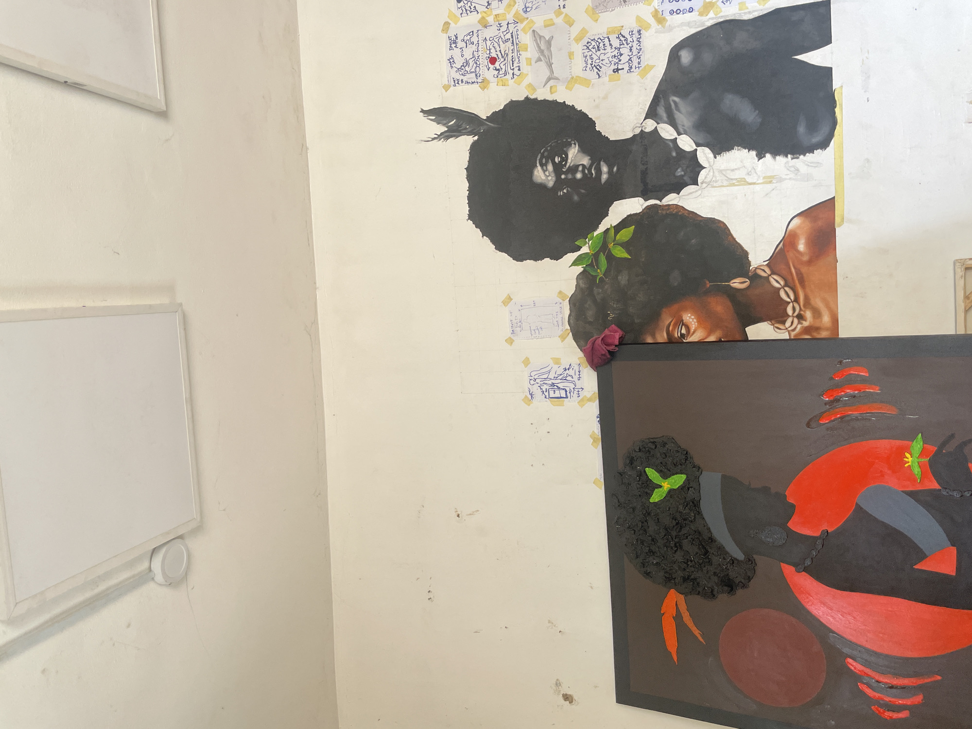 The image shows a section of a wall in what appears to be a room in Ado, Nigeria. The wall is off-white and shows signs of wear and staining. There are two large paintings, one above the other.
The top painting features two people, one above the other, with large afros. The person on top is depicted in black and white, while the person below is in color. Both figures are wearing necklaces. Small pieces of paper with markings are taped to the wall around the top painting.
Below that painting is another painting in a dark brown frame. This painting features abstract shapes and a silhouette of a person with an afro. The colors used are red, black, brown, and gray, with green accents.
To the left, there are two whiteboards mounted on the wall. A white bottle-like object is attached to the wall below the bottom whiteboard.