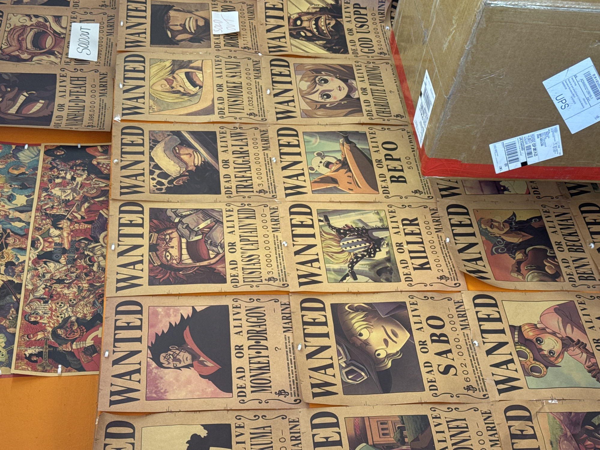 The image shows a collection of One Piece wanted posters laid out on a surface. The posters depict various characters from the series with their names and bounty amounts. A cardboard box with a UPS shipping label is positioned on top of some of the posters. The location is not identifiable from the image, and there is no indication that the image was taken in Barcelona, Spain.