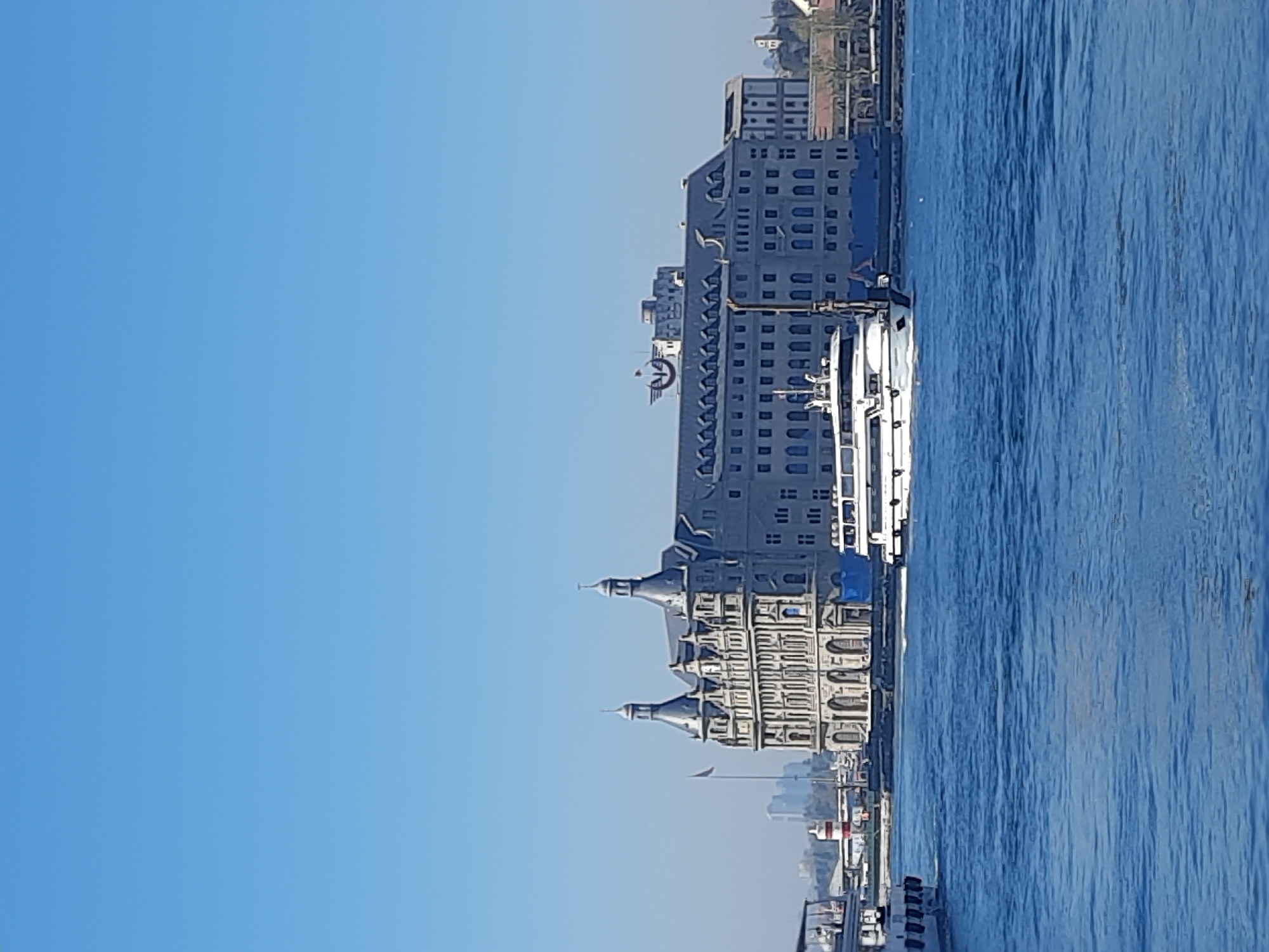The image shows the Haydarpaşa Train Station in Kadıköy, Istanbul, Turkey. The gray, ornate building stands next to a body of water. A ferry is docked at the pier near the station. The sky is a clear blue.