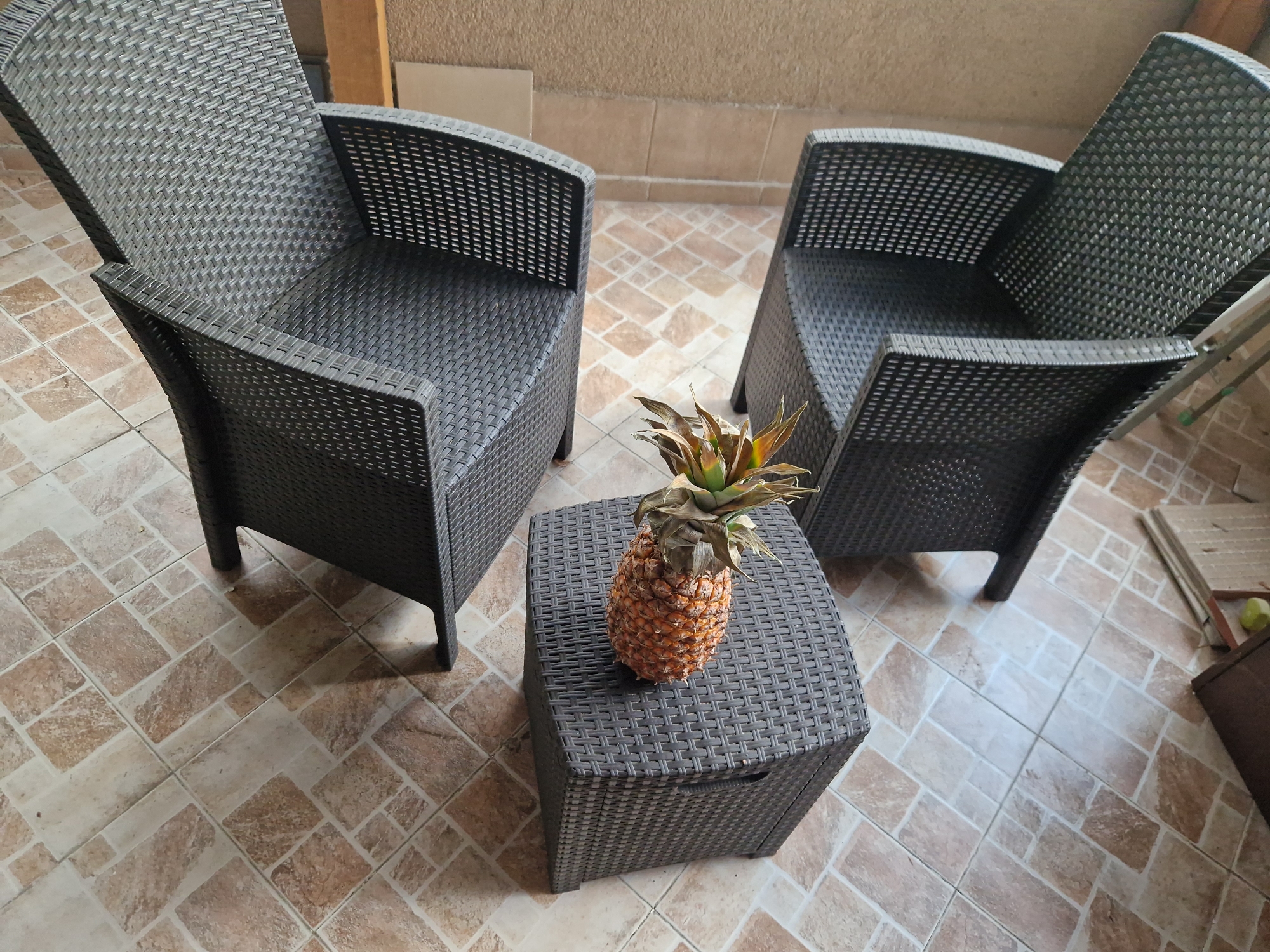 The image shows an outdoor patio setting. Two dark brown wicker armchairs are arranged around a matching wicker side table. A ripe pineapple sits atop the side table. The patio is tiled with a patchwork pattern of beige and brown tiles. The background shows a beige wall. Based on the architectural style, materials, and climate suggested, the location could be a patio in Sofia, Bulgaria. No people are visible.