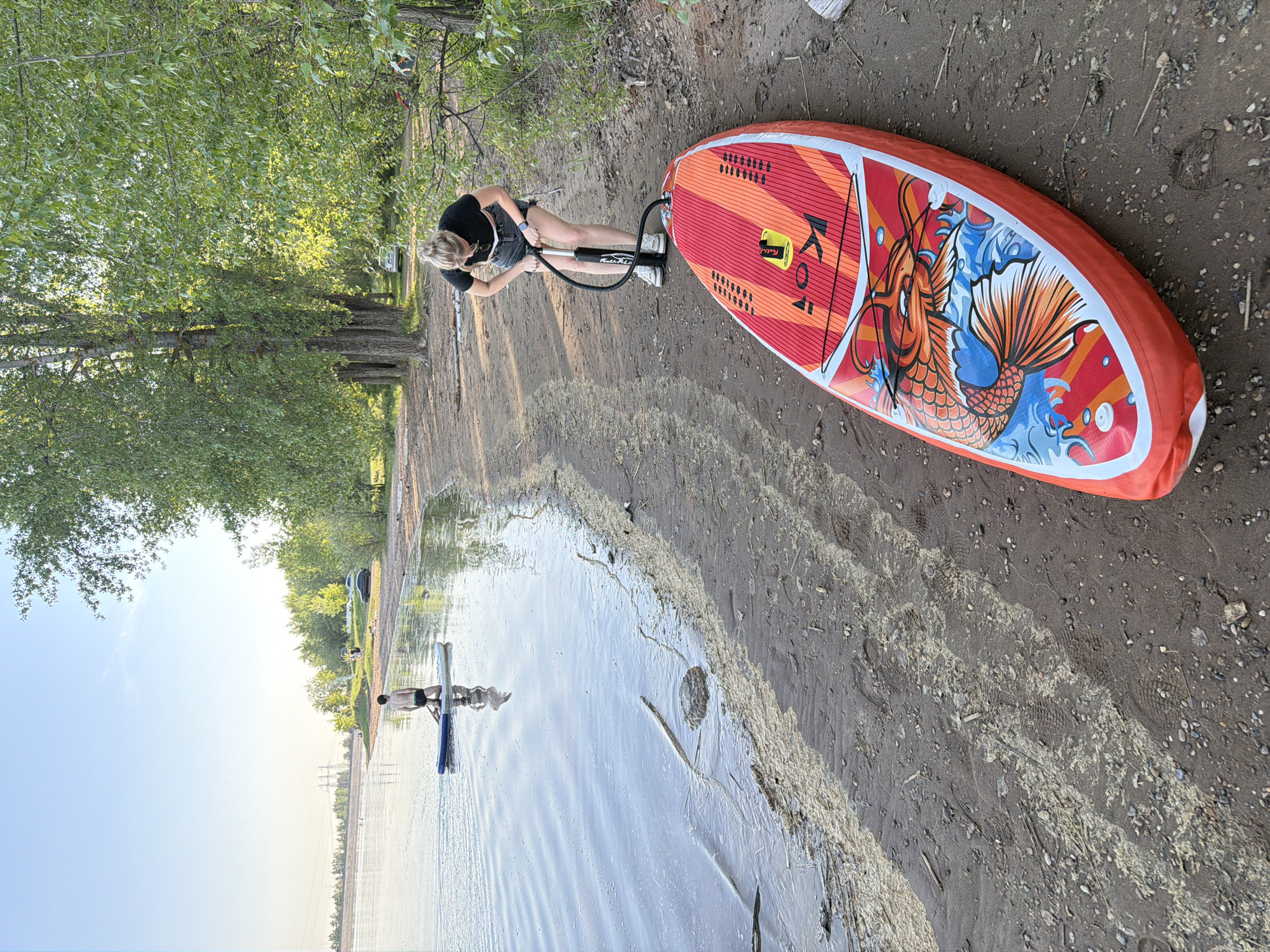 A young woman with blonde hair pulled into a bun is inflating a red, orange, and white inflatable paddleboard with a koi fish design on a sandy beach. She is wearing a black shirt and dark shorts. The paddleboard is connected to a hand pump via a black hose. The beach has scattered pebbles and leads to a body of water. In the water, a person is standing on another paddleboard, holding a paddle. Trees line the edge of the beach, and in the distance, power lines and more trees are visible. The photograph was taken in Noviy, Russia.