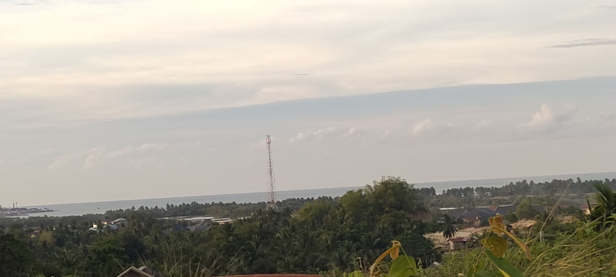 The image presents a panoramic view from a high vantage point in Lhokseumawe, Indonesia, overlooking a coastal area. The sky is overcast with a layer of clouds. A coastline is visible in the mid-ground, bordering a body of water, likely the ocean. A cellular transmission tower is visible in the distance. In the foreground, lush green vegetation dominates, including trees and foliage. Residential buildings are scattered among the trees. A glimpse of the city with what appears to be two smokestacks at the far left side. A part of an unidentified structure is visible in the bottom left. Some yellow flowering plants are in the bottom right corner.