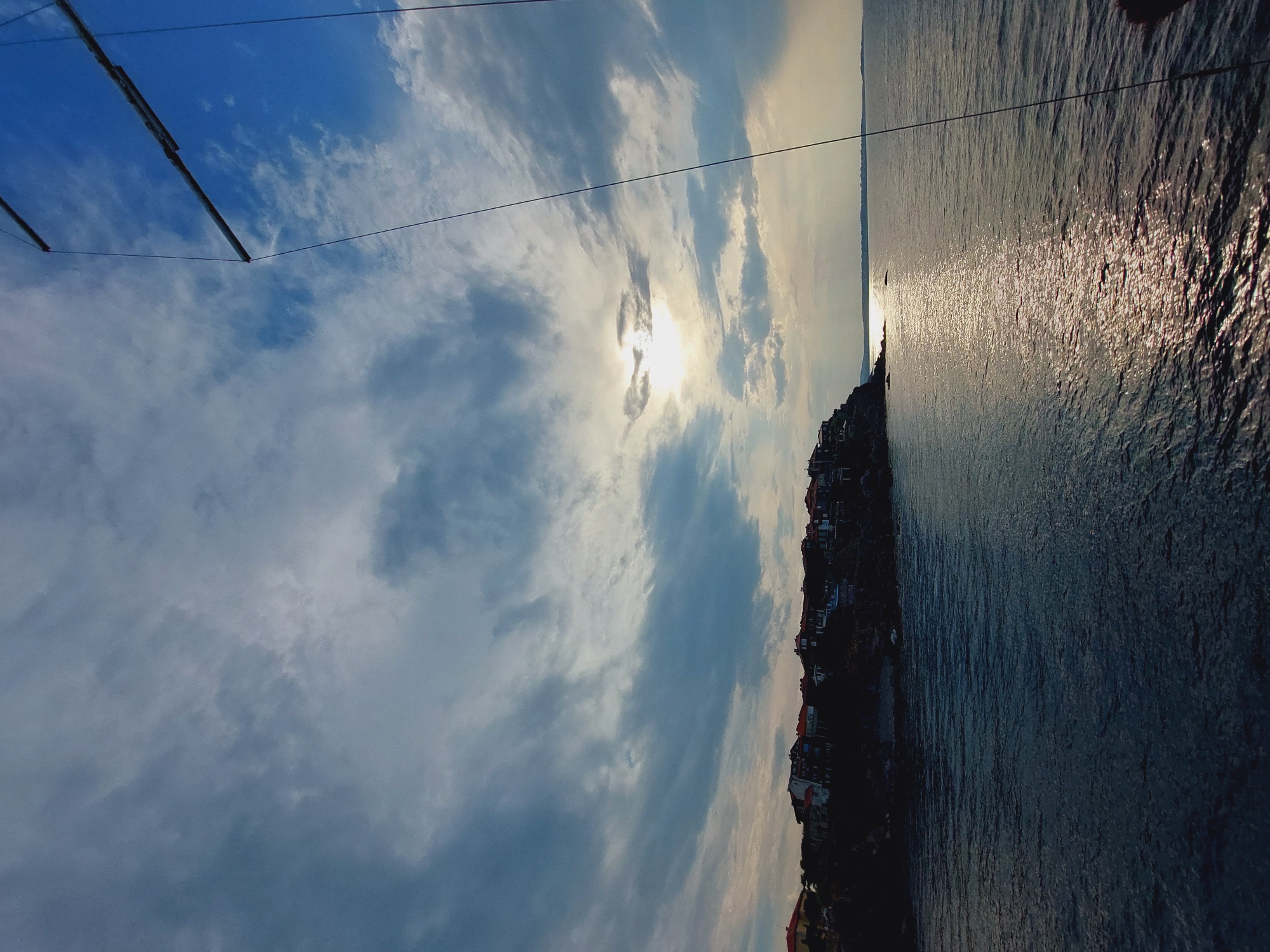 This is a wide-angle, eye-level shot of the coast of Sozopol, Bulgaria, as viewed from the water. The sun is setting or rising, casting a golden reflection on the sea. The sky is filled with dramatic clouds, with patches of blue visible. On the left side of the frame, a rocky cliff juts out into the sea, topped with a dense cluster of red-roofed buildings, characteristic of Sozopol's old town. The water is dark and rippled. A mast and rigging of a sailboat are visible in the upper right corner of the image, suggesting the perspective of someone on a boat. There are no people visible in the scene.