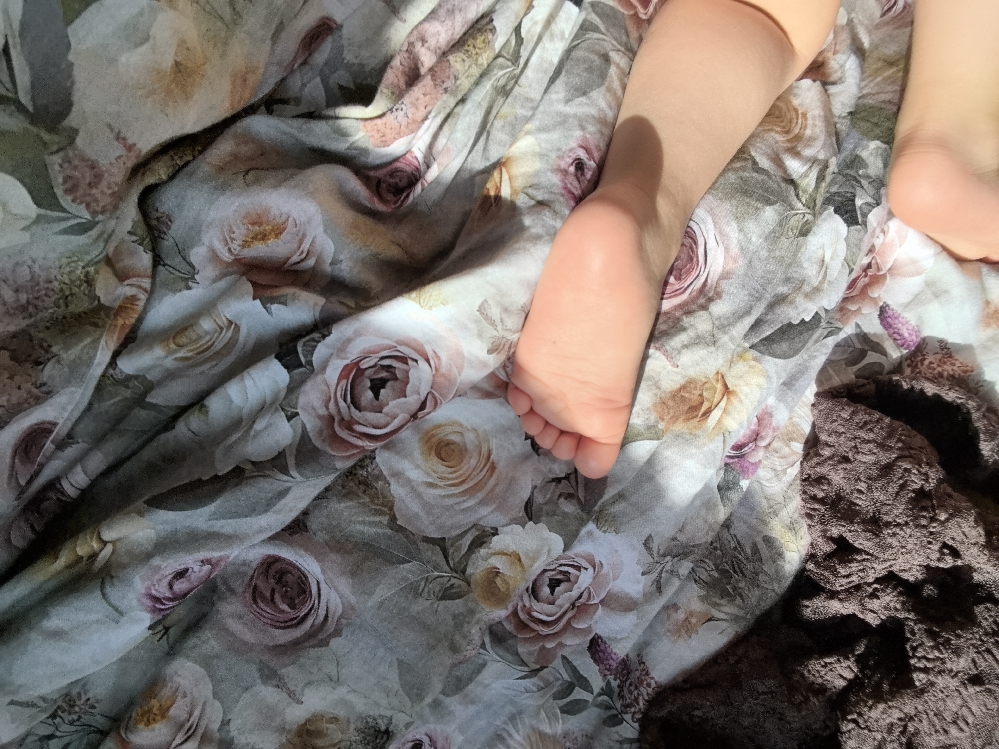 The image shows a close-up view of a bed with a floral patterned sheet. On the sheet, there are two human legs. The bottom of one foot is visible in the left center of the image. The other foot is partially visible in the upper right corner. There is also a dark brown textured blanket or fabric in the lower right corner of the image. The photo was taken indoors under natural light. There is no visible indication of the location being Stara Zagora, Bulgaria.