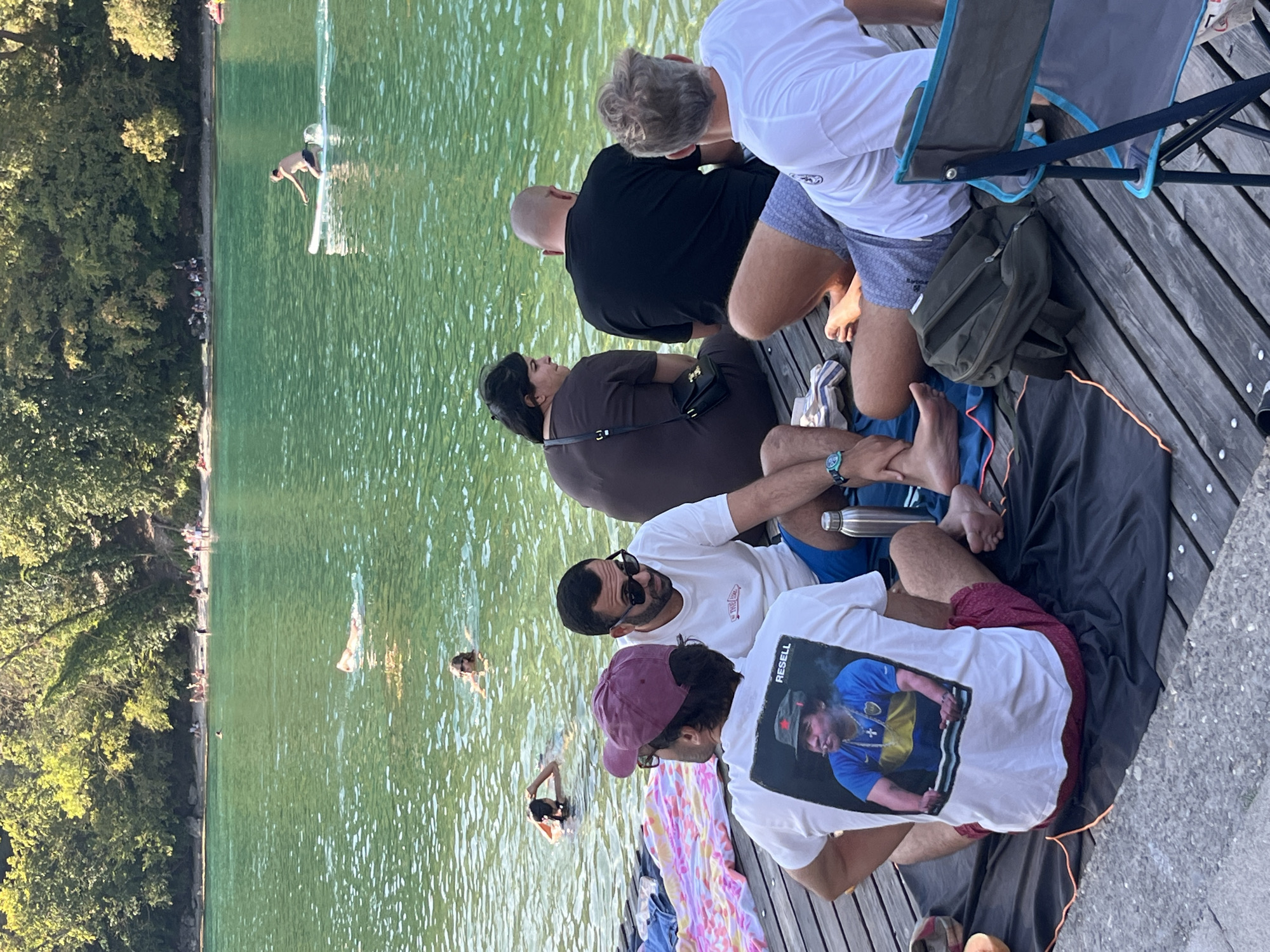 In Geneva, Switzerland, on a wooden pier extending over a large body of water, a group of individuals are gathered. The water, a vibrant emerald green, is populated by several swimmers and a person on a paddleboard. In the foreground, four men are seated on the pier. One man, wearing a burgundy baseball cap and glasses, is facing away from the camera, his back adorned with a t-shirt featuring a printed image of Diego Maradona. Next to him, another man in sunglasses and a white t-shirt is looking down. To their right, a bald man in a black t-shirt is also facing away, and further to the right, a man with blonde hair in a white t-shirt is partially visible, facing the water. Towels and a backpack are scattered on the pier. In the background, the shoreline is lined with lush green trees, and a beach area is visible with more people relaxing. The sun is shining, casting a warm glow over the scene, indicative of a summer day.