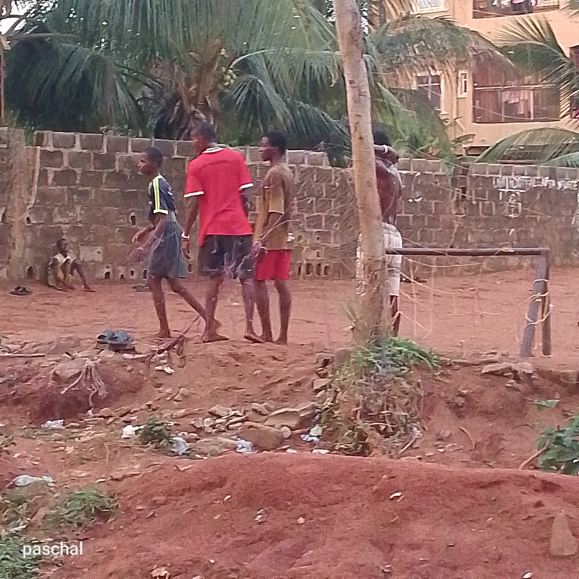 The image depicts a group of boys playing football in Ukwu-Abwa, Nigeria. Four boys are visible in various states of action related to the game. One boy is wearing a red shirt and dark shorts, another a navy and yellow jersey with dark shorts, a third a tan shirt and red shorts, and the fourth boy wears light-colored shorts and is shirtless. A football goal is positioned on the right side of the frame. Another boy is sitting on the ground near a brick wall in the background. The ground is mostly dirt with scattered rocks and vegetation. In the background, a building with multiple stories is visible. Palm trees are also present in the background.
