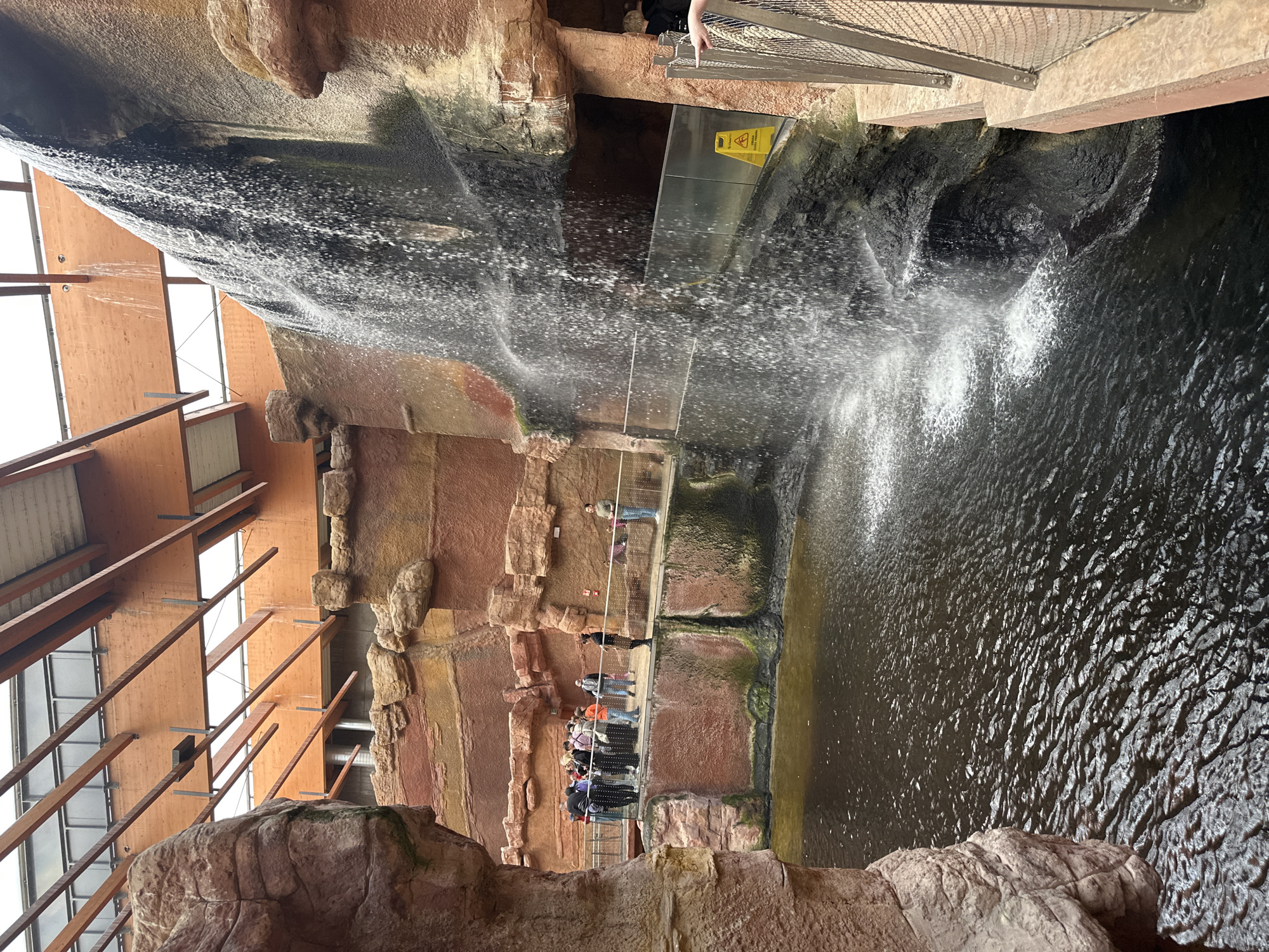 The image shows the inside of the Africarium at the Wrocław Zoo, Poland. A large artificial rock formation with a waterfall dominates the scene. A group of people is standing on a suspended walkway or bridge, viewing the waterfall and the water below. The water appears dark and turbulent. The architectural structure of the building is visible with wooden beams supporting a skylight or roof. Safety signs and metal mesh barriers are present in the upper right corner of the image. The artificial rock structure is multi-toned with shades of brown, red, and tan.