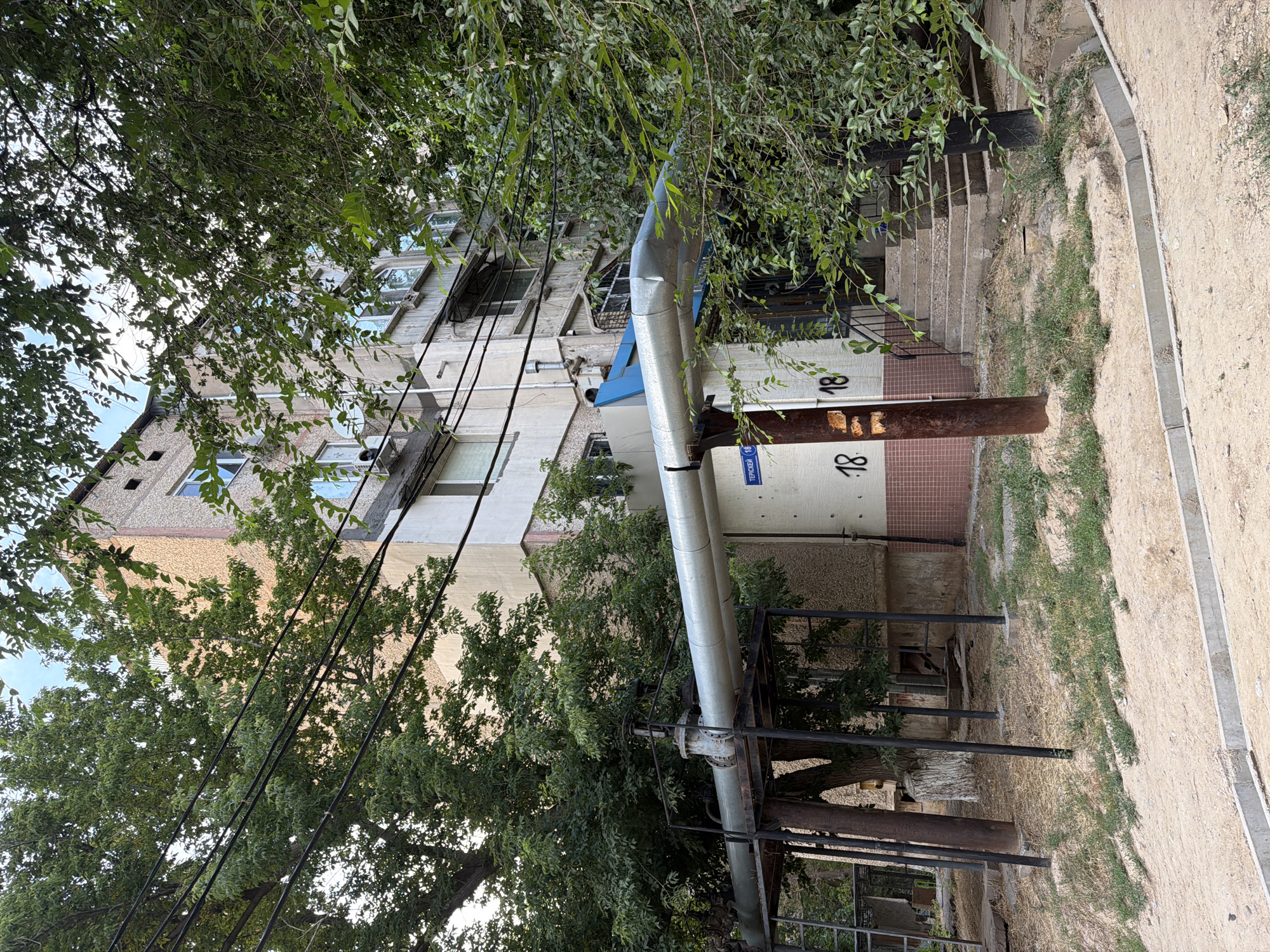 The image depicts an exterior view of a multi-story residential building in Shymkent, Kazakhstan. A large, insulated pipe runs horizontally across the scene, supported by metal structures. The building itself appears to be constructed of concrete and brick, with visible windows and air conditioning units. Green foliage from trees and bushes surrounds the building, partially obscuring it. Street numbering is visible on the building, with "18" clearly marked. A street sign with Cyrillic text, "ТЕРИСКЕН," is also present. The ground in front of the building is a mixture of dirt, sand, and sparse vegetation, with a concrete curb along the foreground. The sky is visible through the tree branches, indicating an outdoor, daytime setting. There are no people visible in the image.