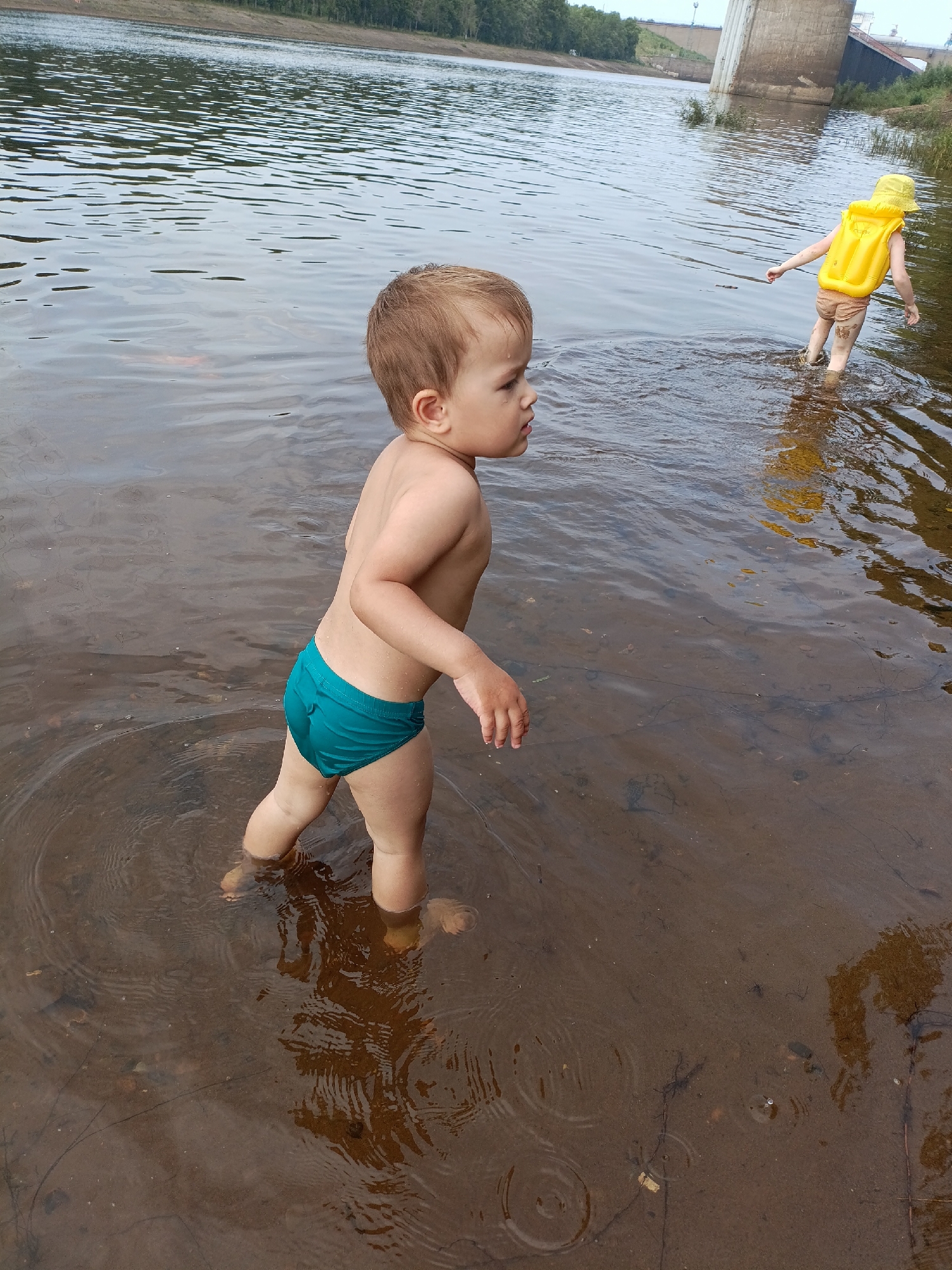 In a shallow body of water with a muddy bottom, two young children are playing. In the foreground, a toddler wearing turquoise swim trunks stands facing away from the camera, with their body angled to the right. Their head is turned to the right, and they appear to be looking towards the other child. In the background, another child, wearing a yellow life vest and a yellow sun hat, stands facing away from the camera, also angled to the right. The water's surface is rippled, and the lighting suggests an overcast or late afternoon day. In the distance, a large concrete structure, possibly a bridge or dam, and a treeline are visible.