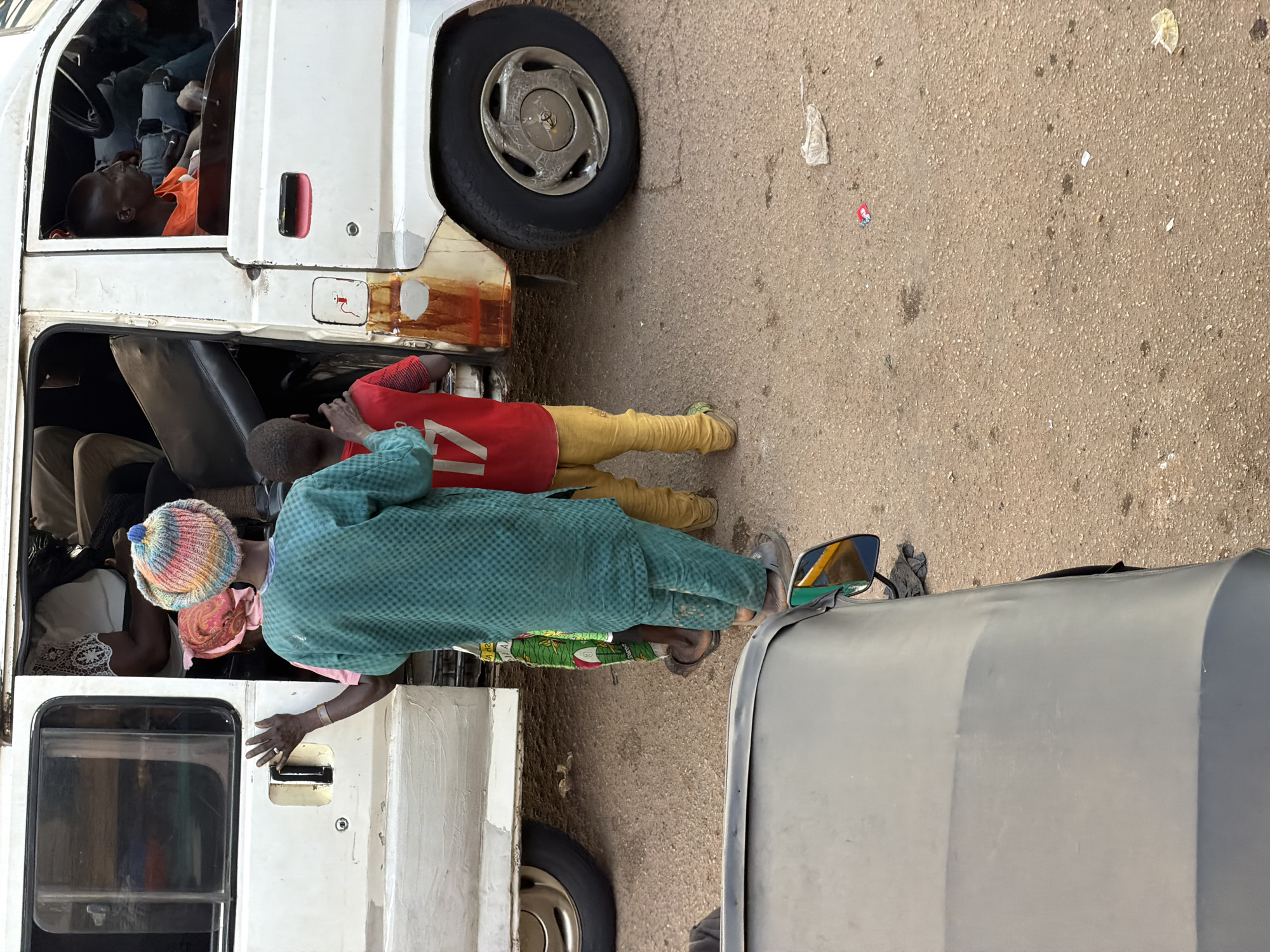 In Bukuru, Nigeria, a white passenger van, likely a commercial vehicle for local transport, is parked on a dusty, light brown road. The van's side door is open, revealing several passengers inside. A person wearing a vibrant, multi-colored knitted hat and a green checkered long-sleeved shirt stands by the open door, facing away from the camera. This individual's left hand is raised and resting on the van's frame. A young child in a red shirt with the number "17" on it and yellow pants stands to the right of the person in the green shirt, also facing away from the camera. Another child, partially visible in a patterned green and yellow garment, stands between the person in green and the child in red. The road surface shows signs of wear and has some small pieces of debris scattered on it. In the foreground, the dark grey roof and side mirror of another vehicle, possibly a tricycle or auto-rickshaw, are visible, suggesting this scene is captured from the perspective of a vehicle following the van. The overall impression is a busy street scene in Nigeria, likely during daytime given the lighting.
