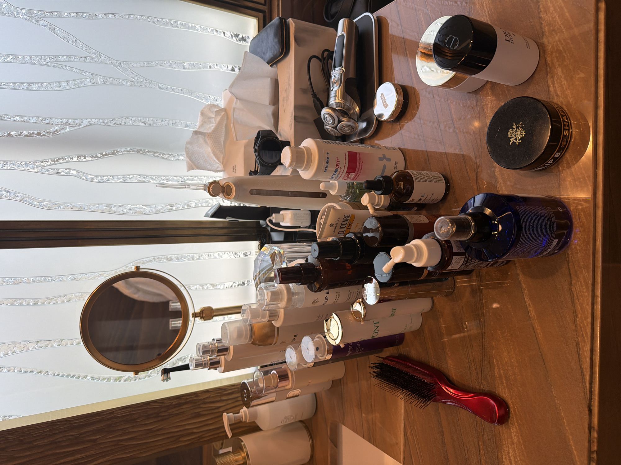 The image depicts a collection of skincare and grooming products arranged on a polished wooden surface, likely a vanity or dresser. The items include various bottles, jars, a hairbrush, a facial steamer, a tissue box, a razor, a mirror, and what appears to be a compact or cream container. The background features a frosted glass panel with an abstract, vertical design and a wooden frame. The lighting suggests an indoor setting, possibly a bathroom or dressing area. There is no indication of the city of Moscow, Russia, or any specific location within it. The scene is static, with no people or discernible actions. The objects are cleanly arranged, suggesting organization and personal care.