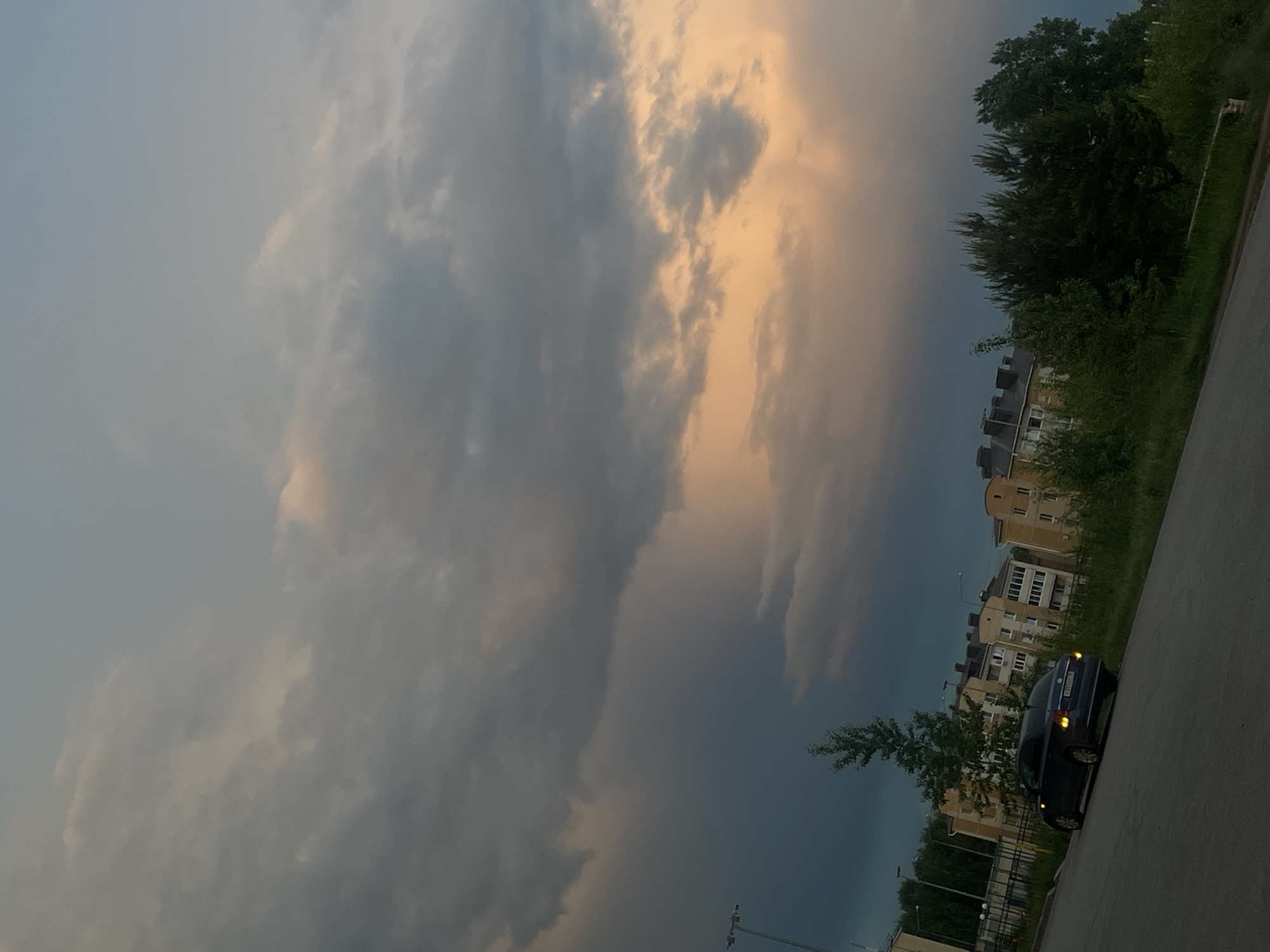 The image, oriented vertically, depicts an outdoor scene at twilight, possibly dusk or dawn, with a dramatic sky dominating the composition. Heavy, dark grey clouds stretch across most of the sky, with a prominent band of warm orange and yellow light breaking through on the right side, suggesting either the sun setting or rising, or light illuminating clouds from behind.
On the right side of the frame, a paved road or parking area is visible, alongside which a dark blue or black car (appears to be a sedan) is parked. Its headlights and possibly taillights are visible. The car's license plate shows "A 813 HE" and "116 RUS" (indicating Russia).
Behind the road and car, a row of multi-story apartment buildings with light-colored facades (beige or yellow) and numerous windows can be seen. They are partially obscured by dense green foliage, including tall trees and bushes, which appear dark against the sky. More trees and greenery are scattered throughout the scene.
The lighting creates a moody atmosphere, with the dark clouds contrasting sharply with the bright, warm glow. There are no people visible, and no specific activity is taking place; the scene appears static. The overall impression is that of a quiet moment in a residential area, likely in Votkinsk, Russia, as indicated by the license plate and context.