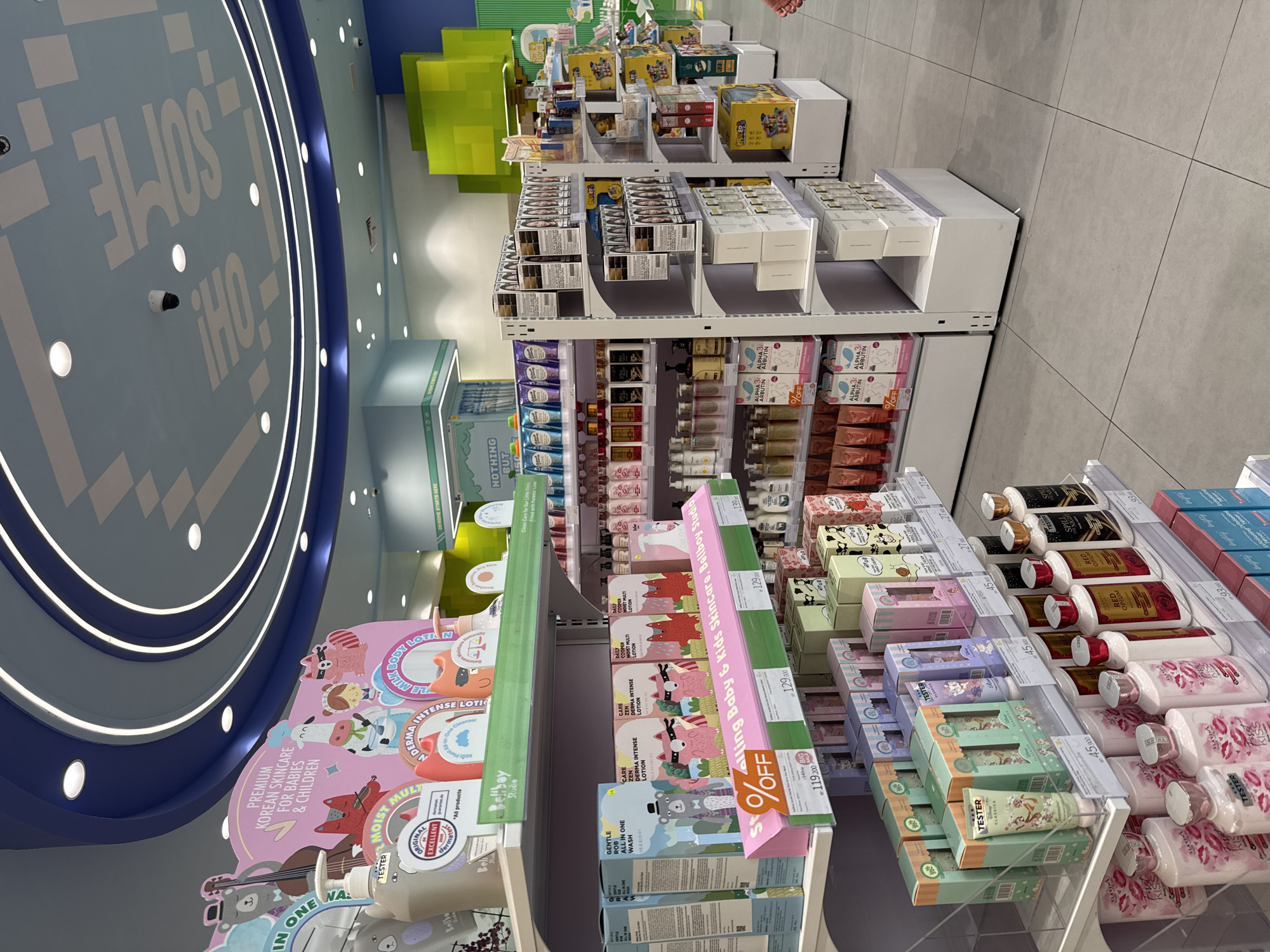 The image shows a well-lit retail space, possibly a beauty or skincare store. The focus is on the product displays, which consist of multiple shelves stocked with various skincare items. The products are diverse, including lotions, creams, and washes, many of which are labeled as being suitable for babies and children. Brightly colored signage and packaging, with some marked as "TESTER." One sign indicates "PREMIUM KOREAN SKINCARE FOR BABIES & CHILDREN" with "Bellboy Studio". There are also sale signs with percentages off. The ceiling features a large circular design with the words "SOME OH!" integrated into the lighting. The floor is tiled and appears clean. No people are visible in the main focus of the image, although a hint of a person is at the top edge of the photo. The image is rotated counterclockwise, indicating it was taken vertically. The location details of Kuta, Indonesia, are not overtly represented in the image and can't be determined from the scene.