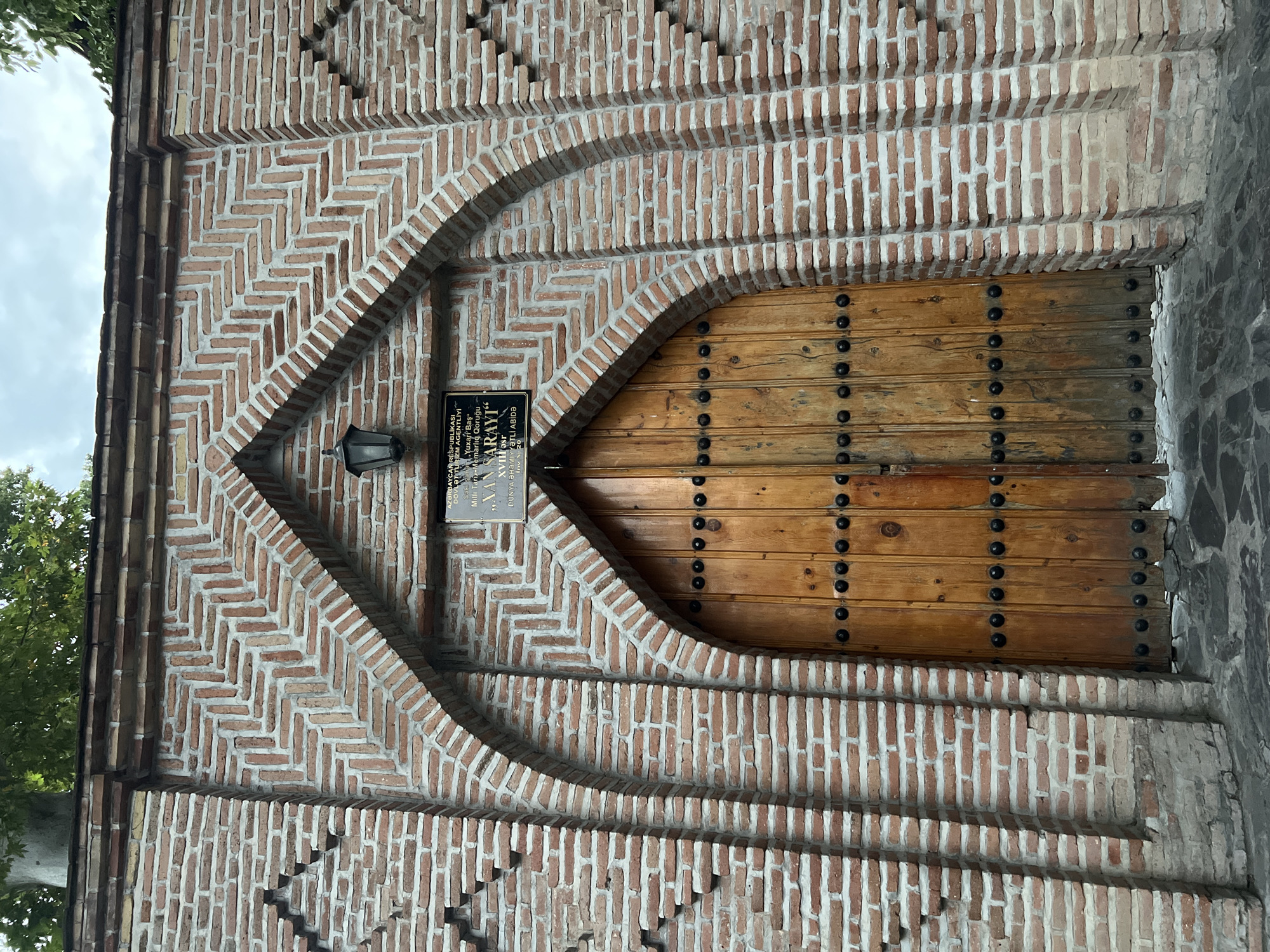 The image shows the entrance to the Palace of Shaki Khans in Shaki, Azerbaijan. The entrance features a large, ornate wooden door with metal studs. The door is framed by a brick archway with intricate herringbone patterns. Above the door, a dark metal lantern is mounted on the brick wall. A plaque with text in Azerbaijani is also visible on the wall. The surrounding brickwork is decorated with geometric patterns, and there are trees visible above the structure, suggesting an outdoor setting.