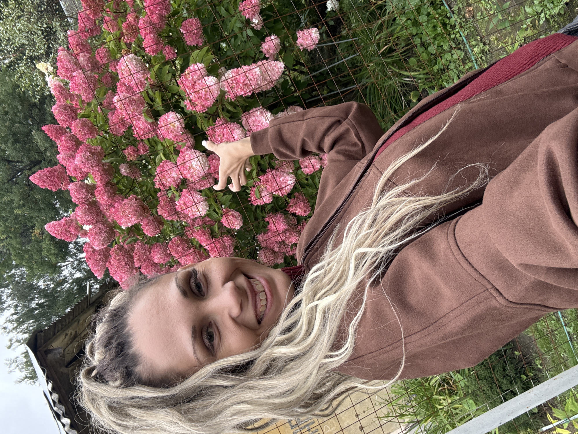 A woman with long blonde hair pulled into a ponytail, wearing a brown zip-up jacket over a red top, smiles at the camera. Her teeth are visible, and she has braces. Her left hand is raised, fingers spread, pointing towards a large bush of pink hydrangeas behind a wire fence. In the background, a small, weathered wooden structure with a corrugated roof is visible, along with green foliage and trees. The scene appears to be outdoors, possibly in a garden or rural area, consistent with a location like Rusanovo, Russia.