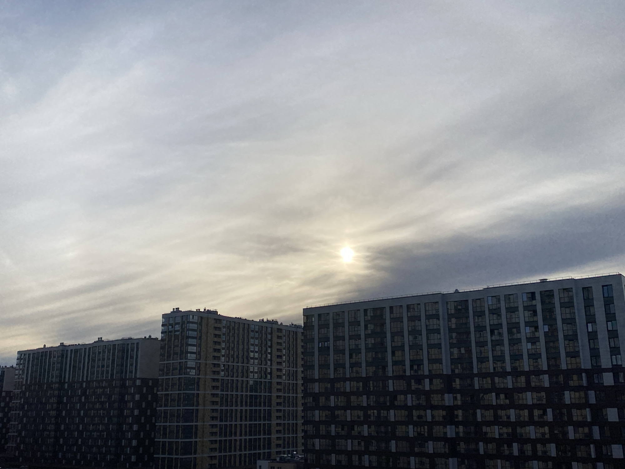 The image depicts a cityscape of Saint Petersburg, Russia, under a partly cloudy sky. The sun is visible, obscured by a layer of clouds. Three multi-story apartment buildings dominate the lower part of the frame. The buildings are characterized by their rectangular shapes and repetitive window patterns. The sky above is filled with elongated, wispy clouds. The light suggests it is either sunrise or sunset. The overall color palette is muted, with shades of gray, white, and brown. There are no visible people, interactions, or actions taking place in the scene.