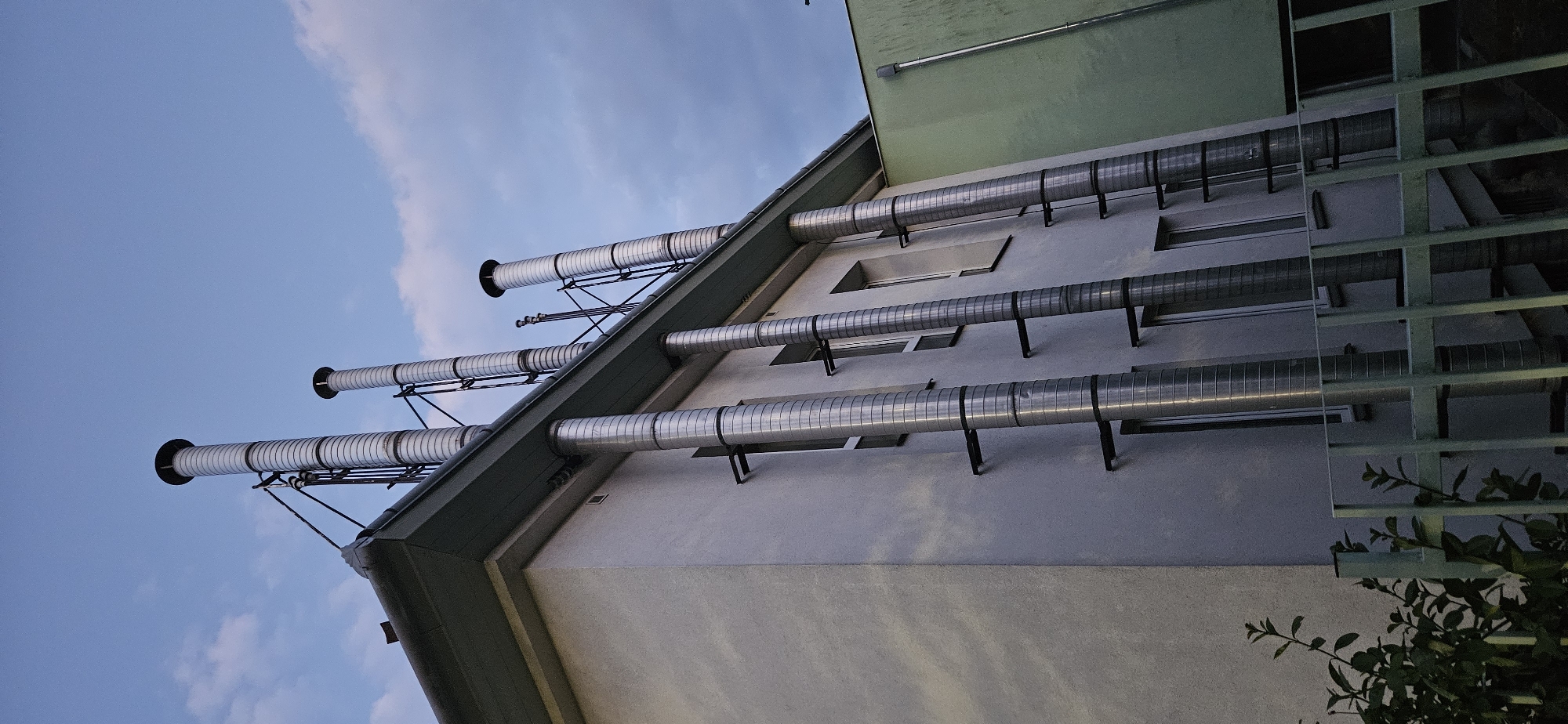 This image captures a low-angle exterior view of a building's ventilation system, featuring multiple large, corrugated metal chimneys extending vertically from the rooftop. The building itself appears to be a light-colored, possibly stuccoed structure with several visible windows. Attached to the side of the building are metal brackets supporting the prominent chimneys, which are staggered in height and position. The chimneys have a ribbed texture due to their corrugated construction. In the background, a portion of an adjacent building, painted a light green, is visible, also with what appears to be a metal pipe running vertically. The sky above is a dusky blue with scattered clouds, suggesting either early morning or late evening. In the foreground, a light green metal fence and some dark green foliage partially obscure the lower section of the building. The scene appears to be an industrial or commercial facility, not a residential one. Based on the context provided, this image is located in Hévíz, Hungary. There are no people or animals visible in the image.