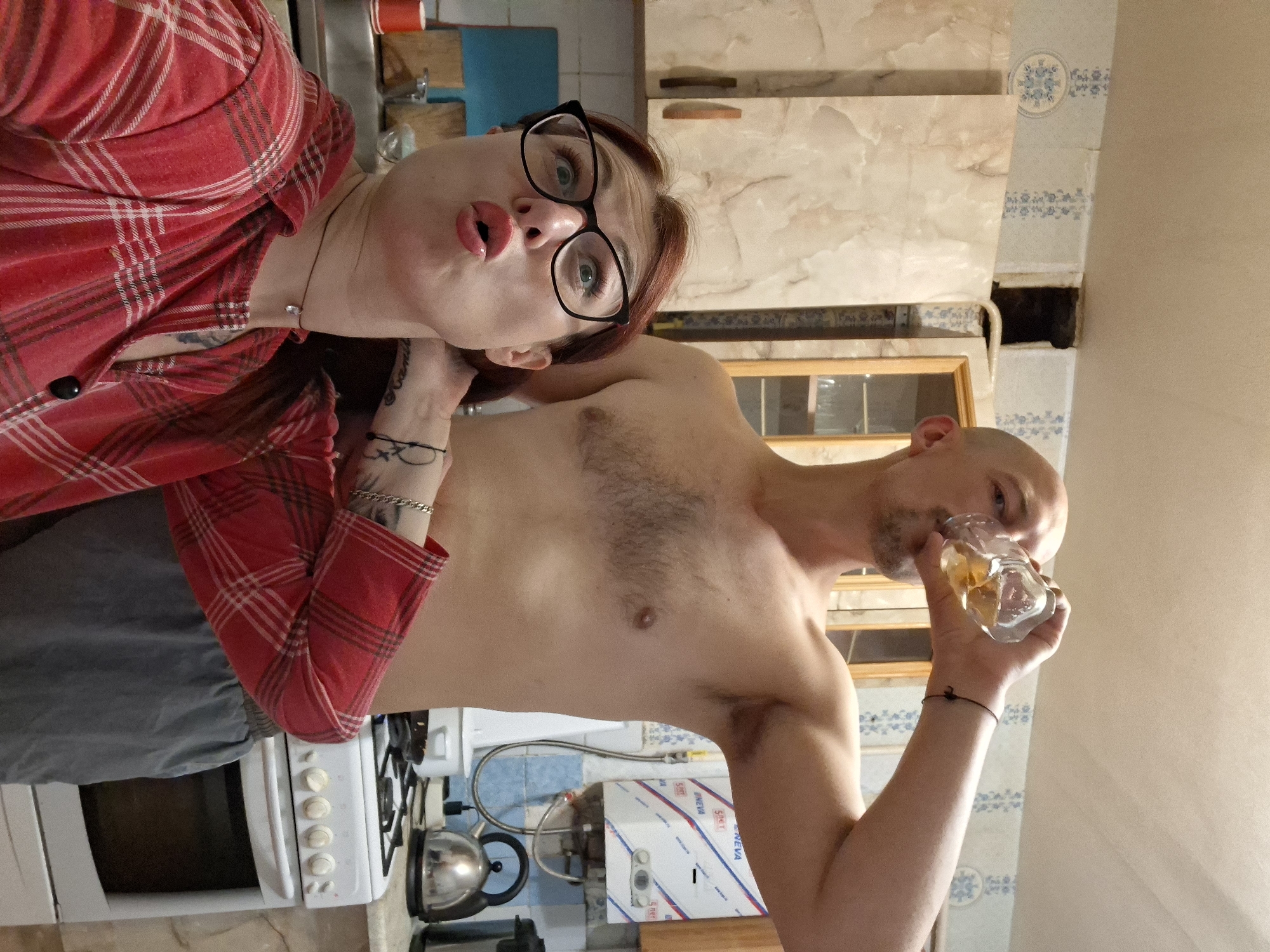 A medium shot shows a couple in a kitchen. The woman in the foreground, with red hair and glasses, makes a surprised expression with her mouth open. She wears a red plaid shirt and a simple necklace. A bald man stands behind her, drinking from a glass of amber liquid. He is shirtless and wears grey shorts. The kitchen appears dated, with patterned wallpaper, light-colored cabinets, and a white gas stove. A water heater is visible on the wall. The setting is likely a private residence in Забалоцкі сельскі Савет, Belarus.