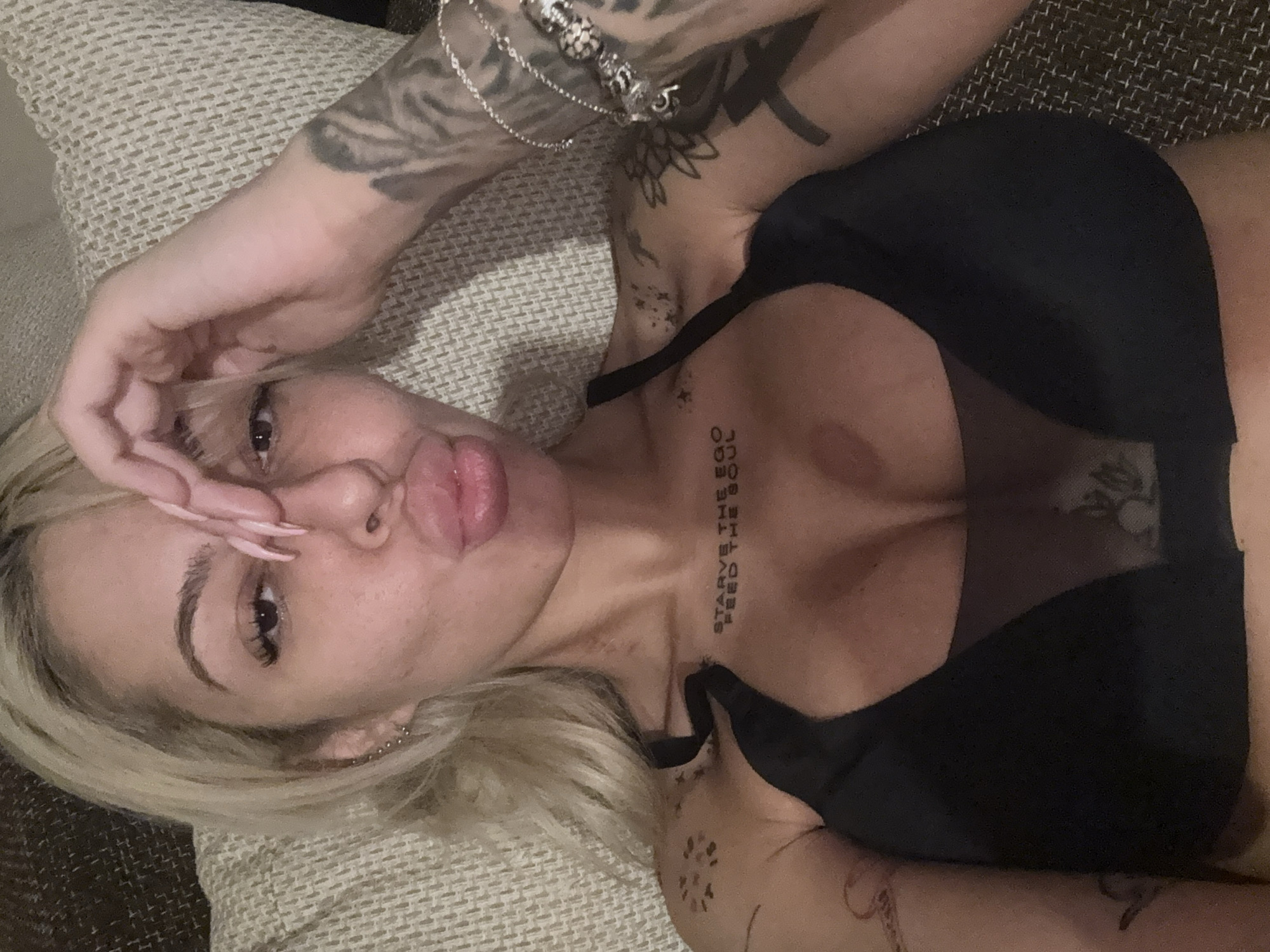 A woman with light blonde hair lies on a textured surface, possibly a cushion or couch. She wears a black mesh bra. Tattoos are visible on her chest, shoulder, and arms, including text that reads "STARVE THE EGO, FEED THE SOUL". She has a silver bracelet on her left wrist and her left hand is near her forehead. She has light-colored nail extensions. The background is indistinct. The location in Plovdiv, Bulgaria is not discernible from the provided image.
