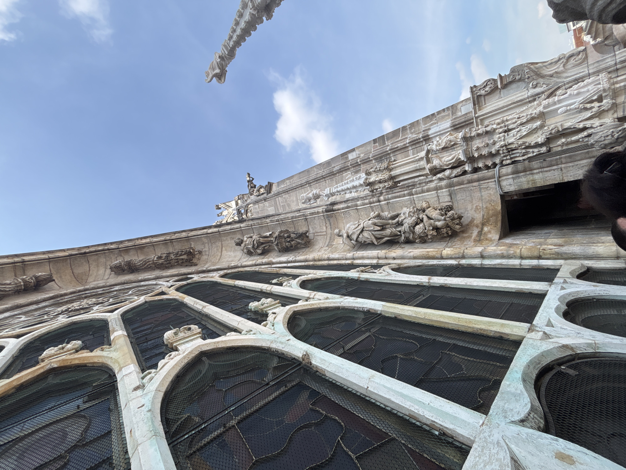 The image shows a low-angle view of the facade of a building in Milan, Italy, likely the Duomo. The architecture features Gothic-style windows with mesh screens and ornate stone carvings, including sculptures of figures and decorative motifs. A gargoyle extends from the upper part of the building against a backdrop of blue sky with scattered clouds. The building's surface is weathered, indicating age. The image's perspective is from ground level, looking upwards at the facade.