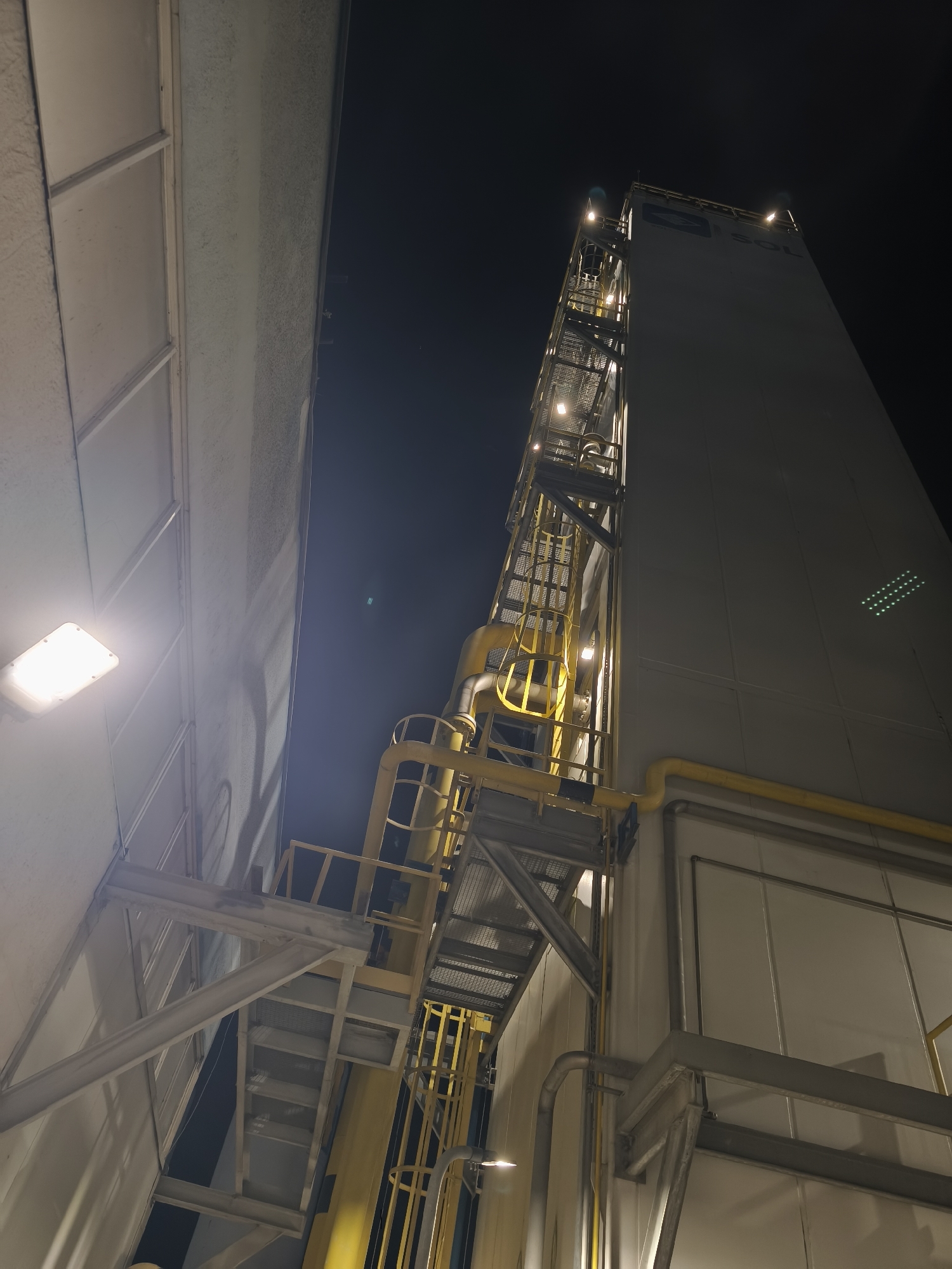 An exterior, low-angle, night time photograph captures a section of a large industrial building. The structure is tall and predominantly white, with a prominent yellow metal staircase and piping system running along its side. The staircase appears to be a multi-level external access route, with lighted platforms and railings. Several bright outdoor lights illuminate the scene, casting harsh shadows and highlights. The building has a section with horizontal paneling, and on its upper right side, a logo with the letters "SOL" and an emblem is visible. The sky is dark, indicating it is nighttime, and faint stars or atmospheric particles are visible. To the left of the tall structure is a lighter colored building with what appear to be large, frosted windows or panels, one of which has a bright rectangular light fixture mounted on it. The overall impression is one of an industrial facility operating at night.