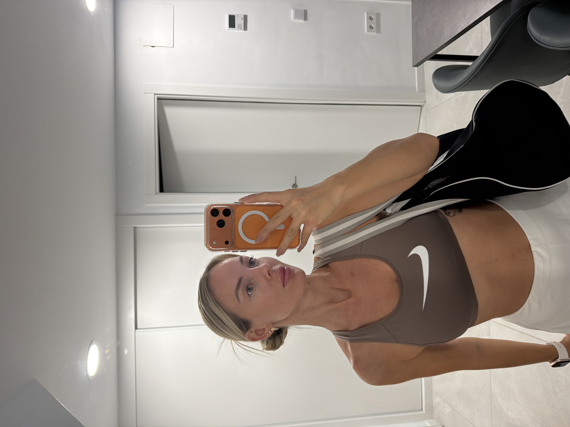 A woman is taking a mirror selfie indoors. She is wearing a brown sports bra and a white skirt. She has blonde hair tied up in a bun and is wearing earrings and a watch. She is holding a phone in her right hand and a black bag over her left shoulder. The background shows a modern interior with white walls and doors, a light-colored tiled floor, and a black chair and table. The scene appears to be in a residential or hotel setting in Finestrat, Spain.