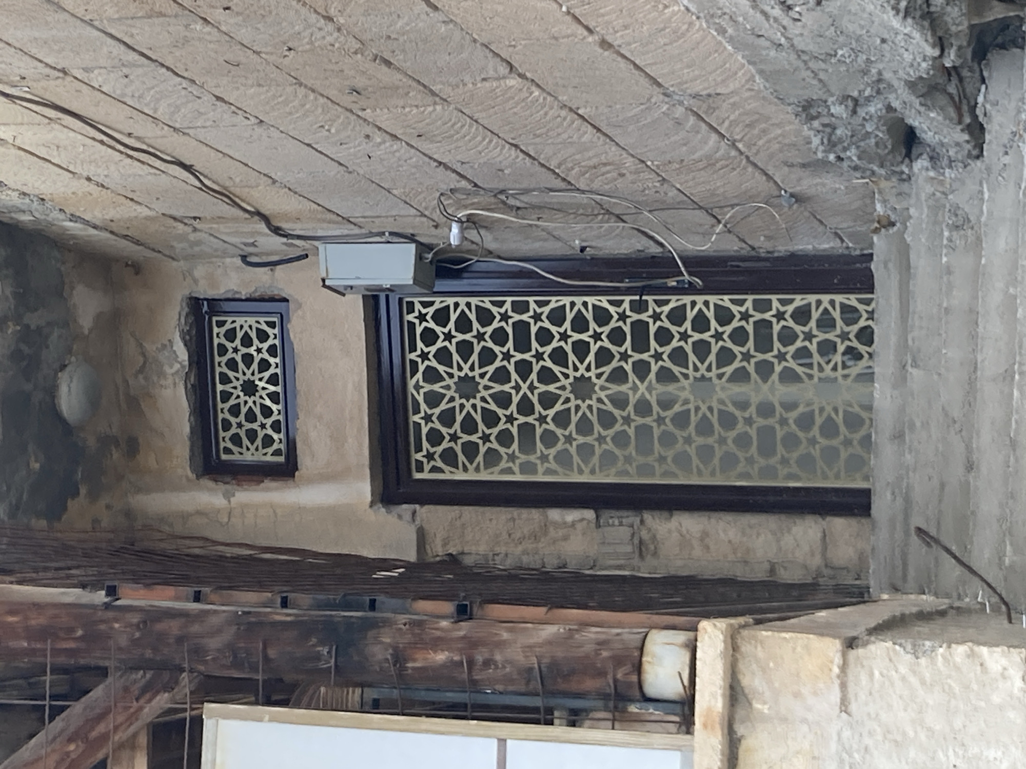 The image depicts a narrow outdoor staircase leading up to a doorway in Avanos İlçe Merkezi, Turkey. The architecture suggests an older building, with a rough-hewn stone wall on the right and a textured, somewhat dilapidated wall on the left. The doorway is framed by a dark brown material, and the door itself features a decorative screen with a geometric star pattern, likely made of metal. Above the door, a small, similarly patterned window is visible. Electrical conduits and a junction box are affixed to the stone wall near the door. The staircase is made of concrete and appears worn. To the left of the stairs, part of a wooden structure with visible metal supports and a white surface is present. The overall scene is one of an urban alleyway, characterized by its aged construction and utilitarian elements. There are no people or animals visible in the frame.