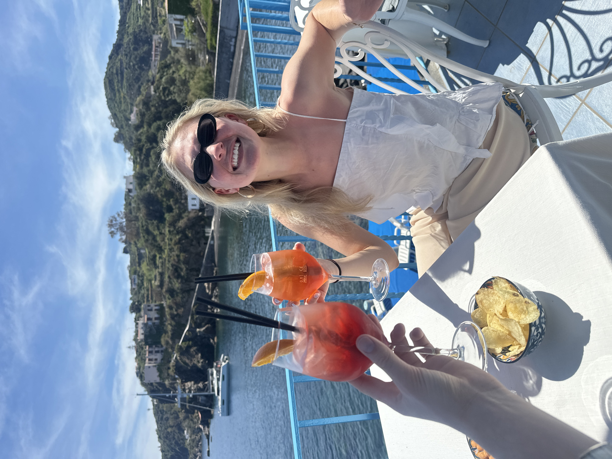 The image portrays a bright and joyful outdoor scene, likely on a restaurant or bar terrace overlooking the sea in Lipari, Italy. A blonde woman, with a wide, radiant smile, is the primary subject. She wears black sunglasses and a white, strapless or spaghetti-strap top, paired with light-colored pants. She holds an orange-hued cocktail, garnished with an orange slice and two black straws, in a stemmed glass, appearing to toast with another person whose hand, holding an identical cocktail, is visible in the foreground.
On the white tablecloth, a small, patterned bowl filled with potato chips is placed next to the drinks. The setting features white outdoor dining chairs with decorative backs and a light blue railing, suggesting a balcony or elevated seating area.
The background offers a picturesque view of a Mediterranean coastal landscape under a brilliant blue sky, dotted with a few wispy white clouds. Steep, verdant hills covered with dense foliage and scattered buildings descend towards a calm body of water, likely the sea, where several boats are moored. The bright sunlight casts distinct shadows, indicating a clear, sunny day, likely in the late morning or afternoon. The woman's cheerful expression and the clinking of glasses evoke a sense of relaxation, celebration, and vacation enjoyment. A small volume marking "0,1l CE" is visible on one of the cocktail glasses.
