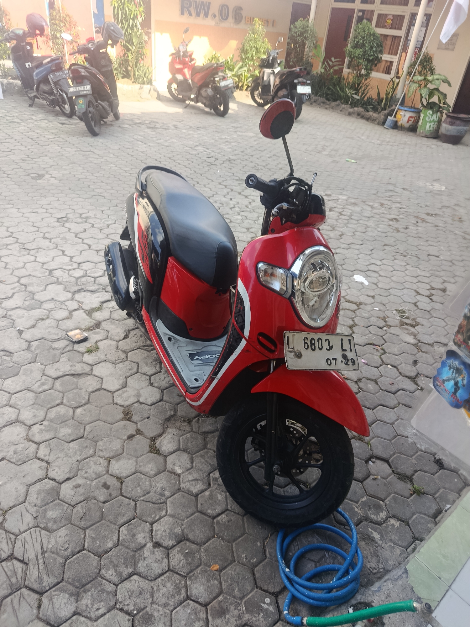 A red and black scooter is parked on a hexagonal paved surface in front of a building. The building has orange and beige walls and several windows. A blue hose with a green nozzle lies on the ground next to the scooter's front wheel. In the background, other motorcycles are parked, and there are plants and a sign with Indonesian text on the building. The image appears to have been taken during daylight.