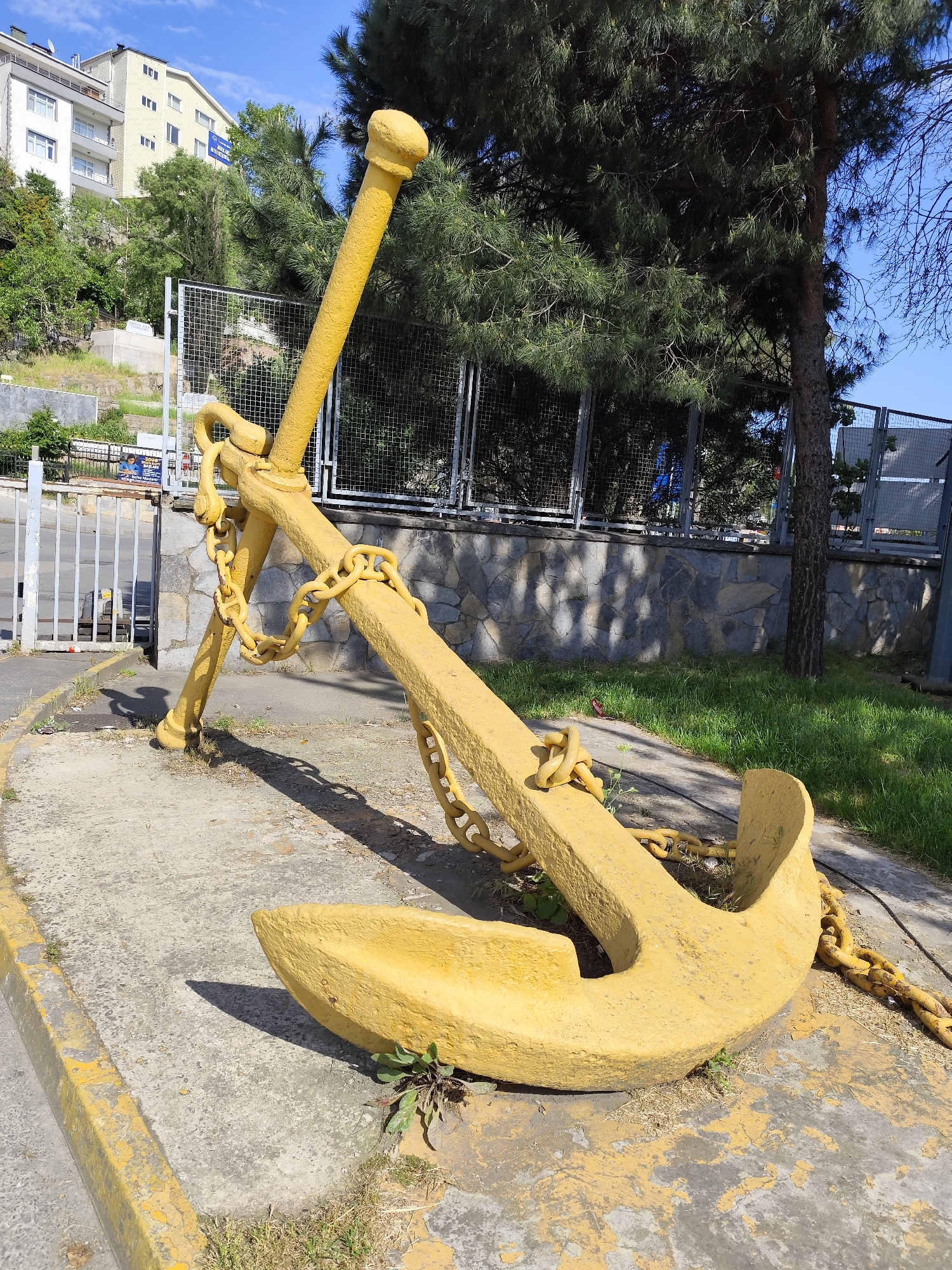 A large, yellow-painted anchor is positioned on a concrete surface with a yellow curb in the city of Unknown, Unknown. The anchor is surrounded by a heavy chain. Behind the anchor is a low retaining wall of rough-cut stone. A fence with a wire mesh top runs atop the stone wall. Beyond the fence is grass and a stand of trees. To the left is a white metal gate, with a building on a hill in the background. The sky is blue with scattered clouds.