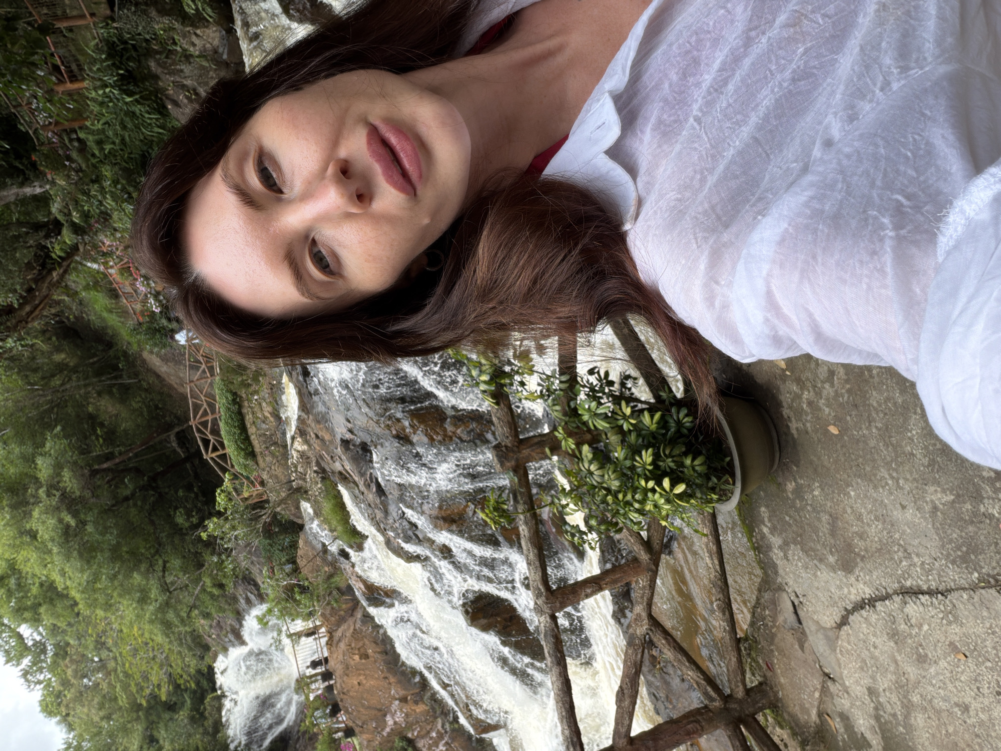 A woman with long, dark hair takes a selfie in front of a cascading waterfall in Da Lat, Vietnam. She is wearing a white, translucent shirt over a red garment. The waterfall is rocky and surrounded by lush green trees and vegetation. A wooden railing runs along a path next to the waterfall. There are potted plants in the foreground.