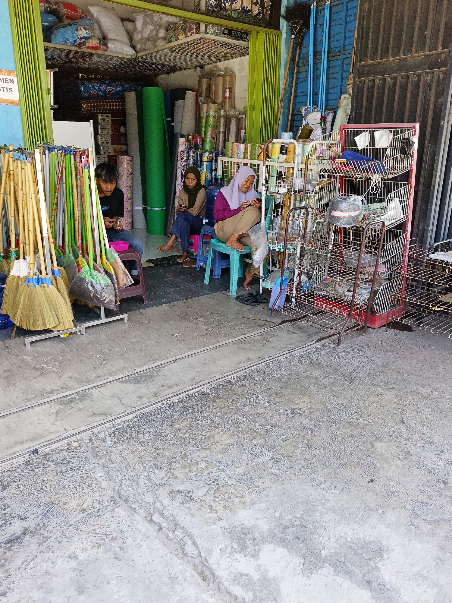 The image depicts the exterior of a small shop in Manisrenggo, Indonesia. In the foreground, a concrete floor with two parallel metal tracks runs across it. To the left, a display of brooms and cleaning tools stands vertically. Behind the brooms, two individuals are seated on small stools. A young male, wearing a black t-shirt, is looking down at his phone. Next to him, a woman wearing a beige hijab and brown clothing is also looking at her phone. Further to the right, another woman, also wearing a hijab and purple top with brown pants, is seated on a blue stool, looking at her phone. Adjacent to the seating area is a multi-tiered metal rack filled with various items, including plastic bags, coils of rope or wire, and other assorted goods. The shop itself is visible in the background, stocked with rolls of carpet or fabric, shelves of goods, and what appear to be cleaning supplies. A blue metal door is visible on the right side of the frame. The overall environment suggests a retail space open to the street, possibly selling household goods or cleaning supplies.