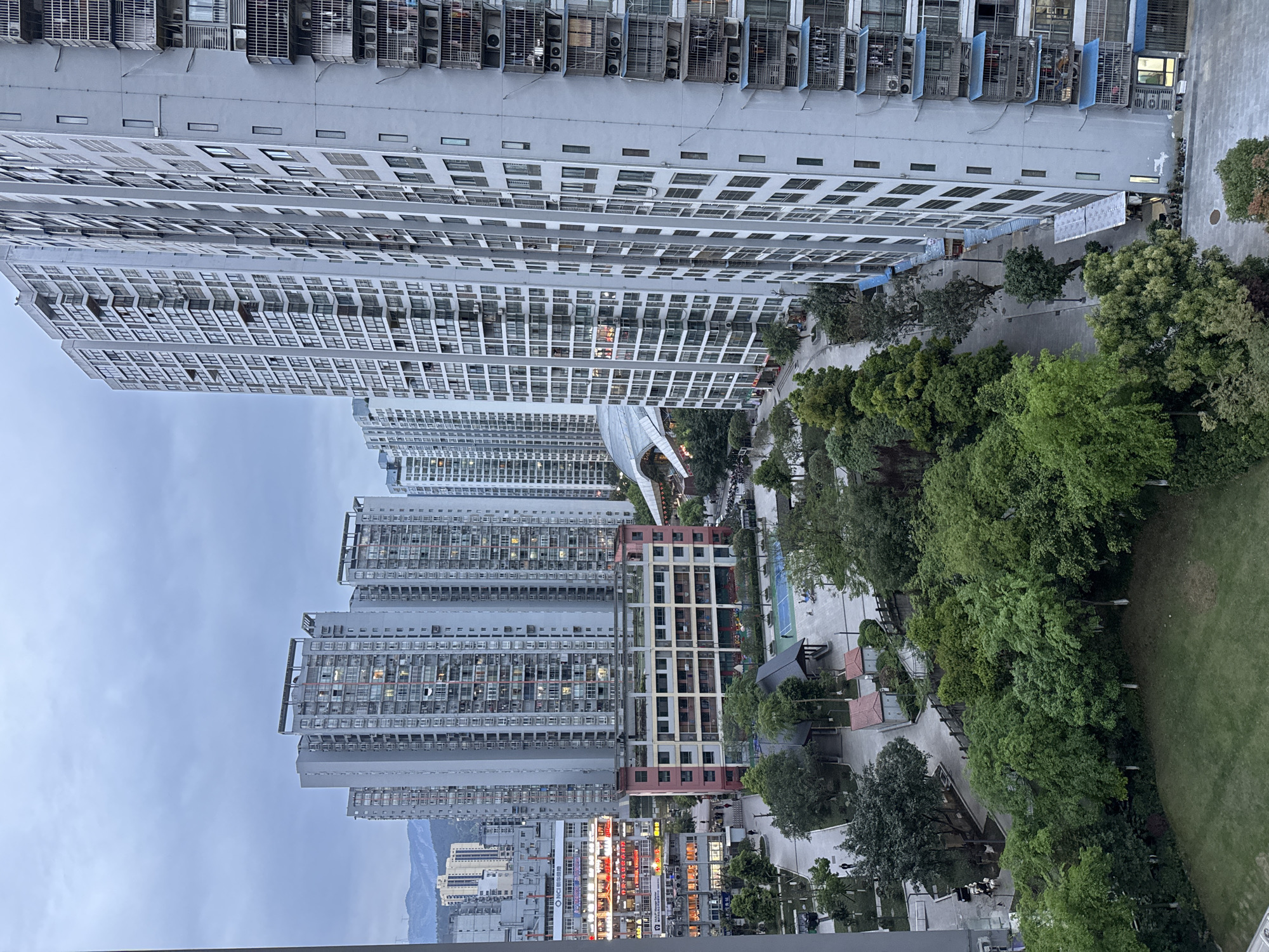 The image shows a high-angle view of an urban environment in Wulong District, China. Multiple tall residential buildings dominate the skyline, characterized by their repetitive window patterns and grey exteriors. Some buildings have air conditioning units protruding from the facades. Pedestrian walkways and green spaces with trees are visible between the buildings. A tennis court and several small structures with red roofs are present in the green space. At the bottom of the image, there are illuminated storefronts and a glimpse of distant mountains. The sky is overcast.
