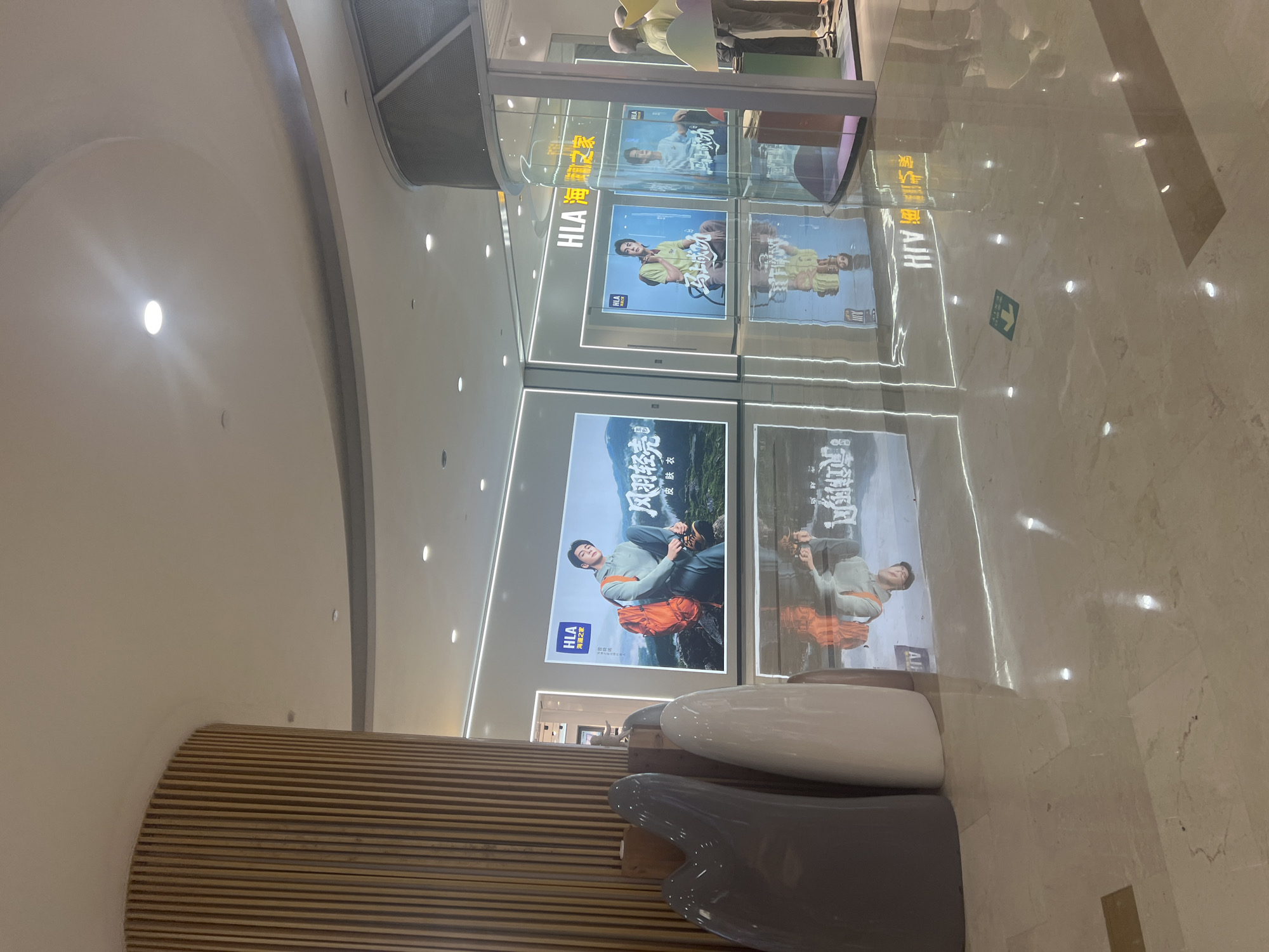 The image depicts the interior of a modern shopping mall, likely in Liandu District, China, with a polished marble floor that reflects the overhead lighting and displays. The scene features a prominent wooden pillar on the left, its vertical slats forming a textured surface. In the background, large illuminated display screens showcase advertisements for the clothing brand HLA. One screen prominently features a man with an orange backpack, posed against a mountainous landscape. Other screens show men in casual attire. To the right, a glass-fronted shop displays mannequins wearing similar clothing. Several abstract, smooth sculptures in shades of gray and white are positioned in the foreground, near the wooden pillar. The overall environment is clean, well-lit, and designed for retail purposes.