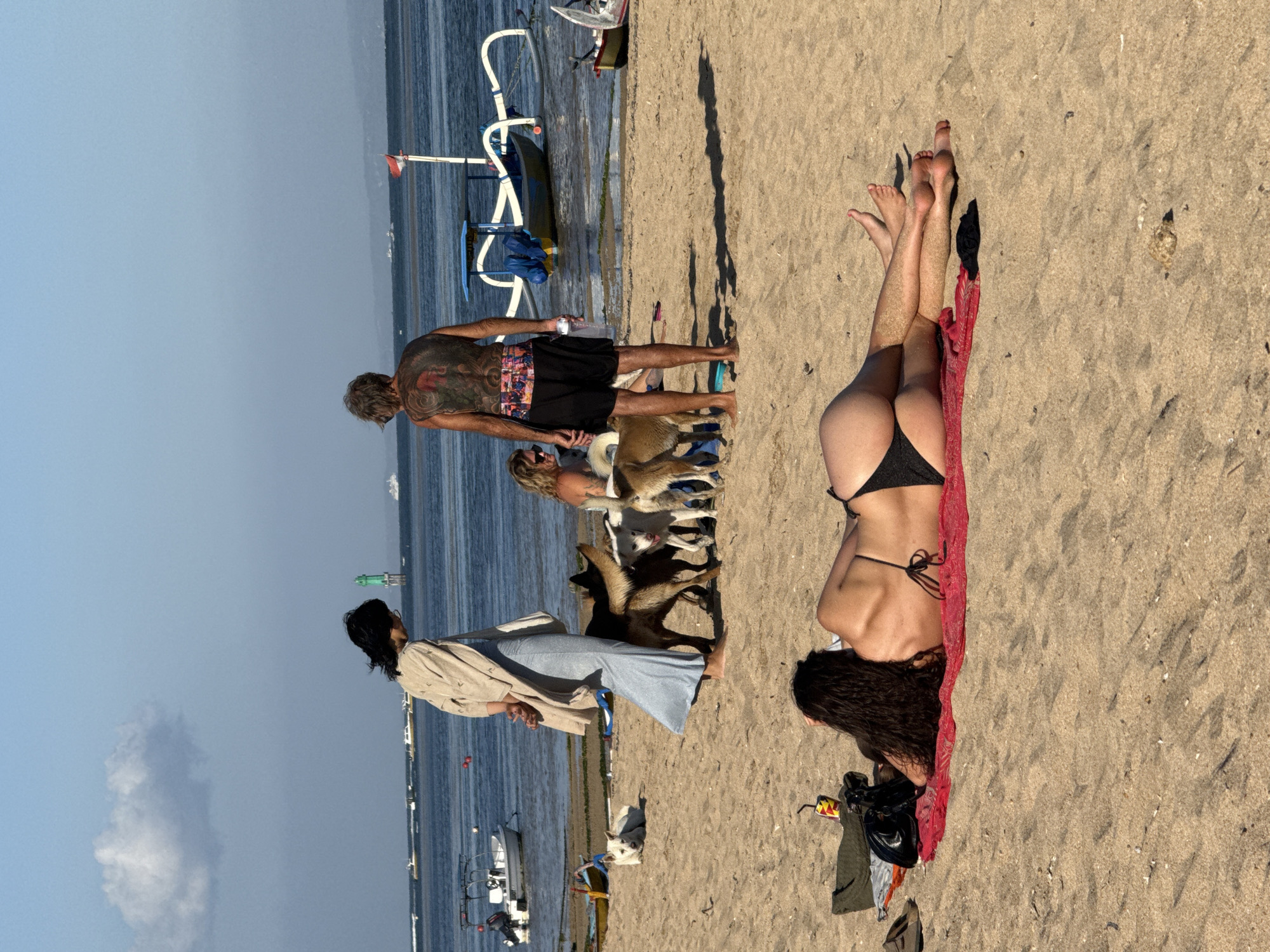 The image depicts a sunlit beach scene with several individuals and dogs near the water's edge. In the foreground, a person lies on a red towel on the sand, wearing a black bikini. To their left, a black bag and a cup with a straw are visible. Further back, a woman in a light-colored cardigan and a long blue dress stands, facing away from the viewer, while several dogs gather around her. Beside her, a man with a large tattoo covering his back, wearing black shorts, stands facing the water. Behind them, a woman with blonde hair is seated. In the background, the ocean stretches to the horizon, dotted with several boats and a green lighthouse. The sand is light brown and appears to be composed of fine grains with some shell fragments. The sky is blue with a few scattered clouds. The scene suggests a relaxed atmosphere on a beach, likely in Denpasar, Indonesia, given the presence of traditional Balinese outrigger boats and the general coastal environment.