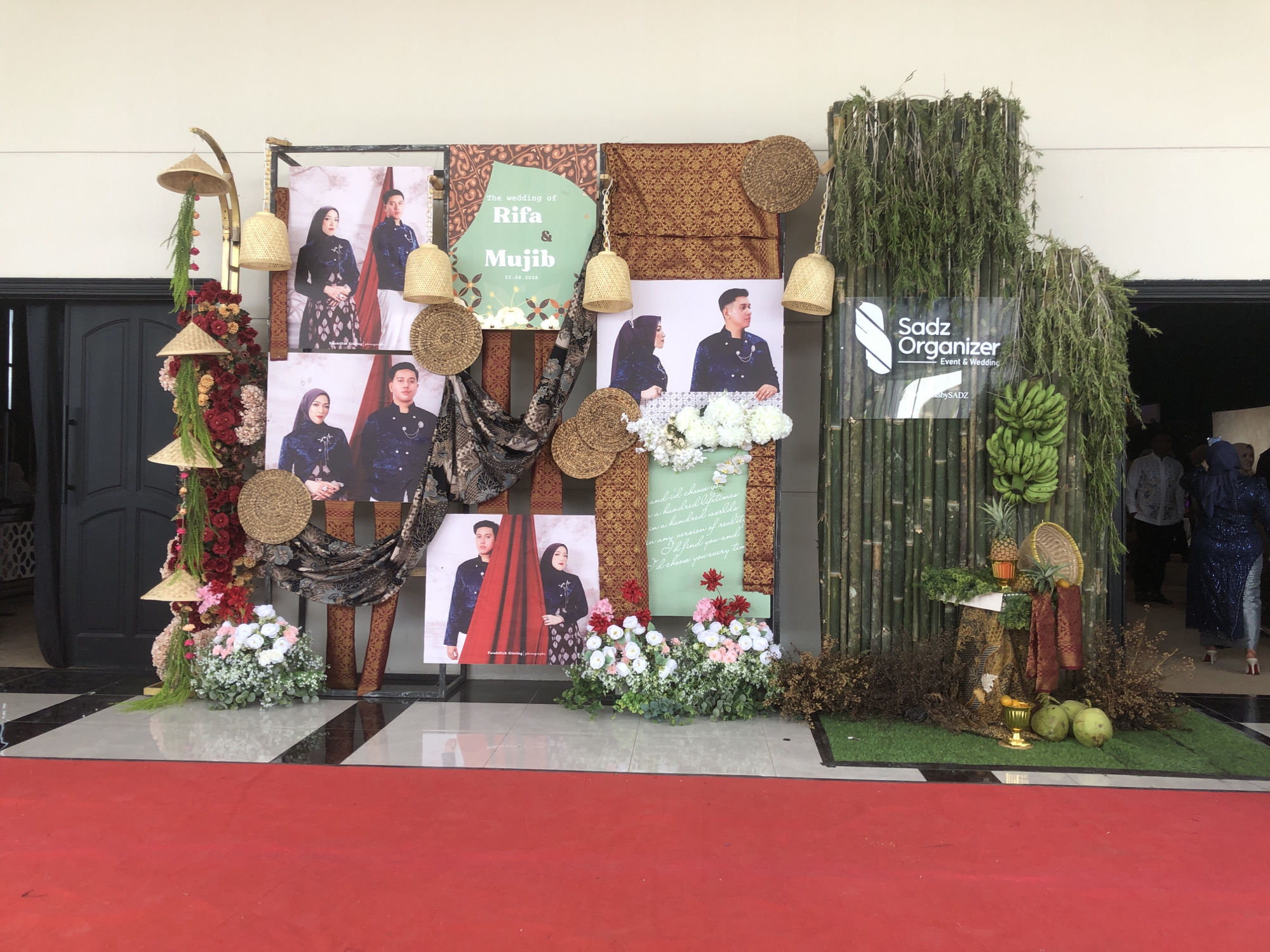 The image shows a wedding backdrop decorated with photos of a couple, flowers, traditional Indonesian fabrics, and bamboo. There is also a sign for "Sadz Organizer Event & Wedding." On the right side, a woman in a blue sequined dress and another person in a grey shirt are visible. The backdrop is set up in front of a building entrance with large black doors and a red carpet leading away from it. The scene appears to be an outdoor event space, possibly in Lhokseumawe, Indonesia, given the traditional elements and the mention of an organizer.