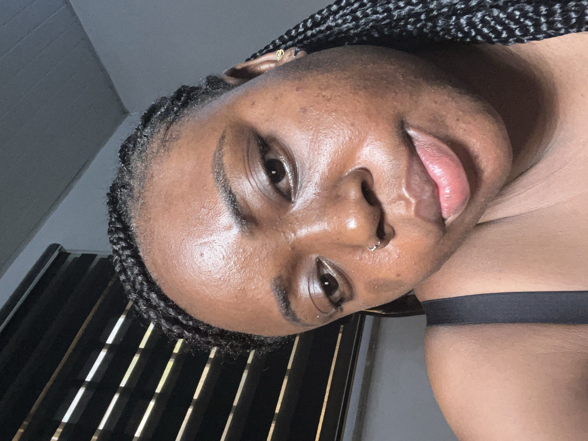 A close-up, angled portrait shows a dark-skinned woman with braided hair. A small gold earring is visible in her left ear. She has a nostril piercing in her left nostril. A black strap crosses her shoulder. A window with blinds is partially visible in the background. No information indicates the location is Abuja, Nigeria.