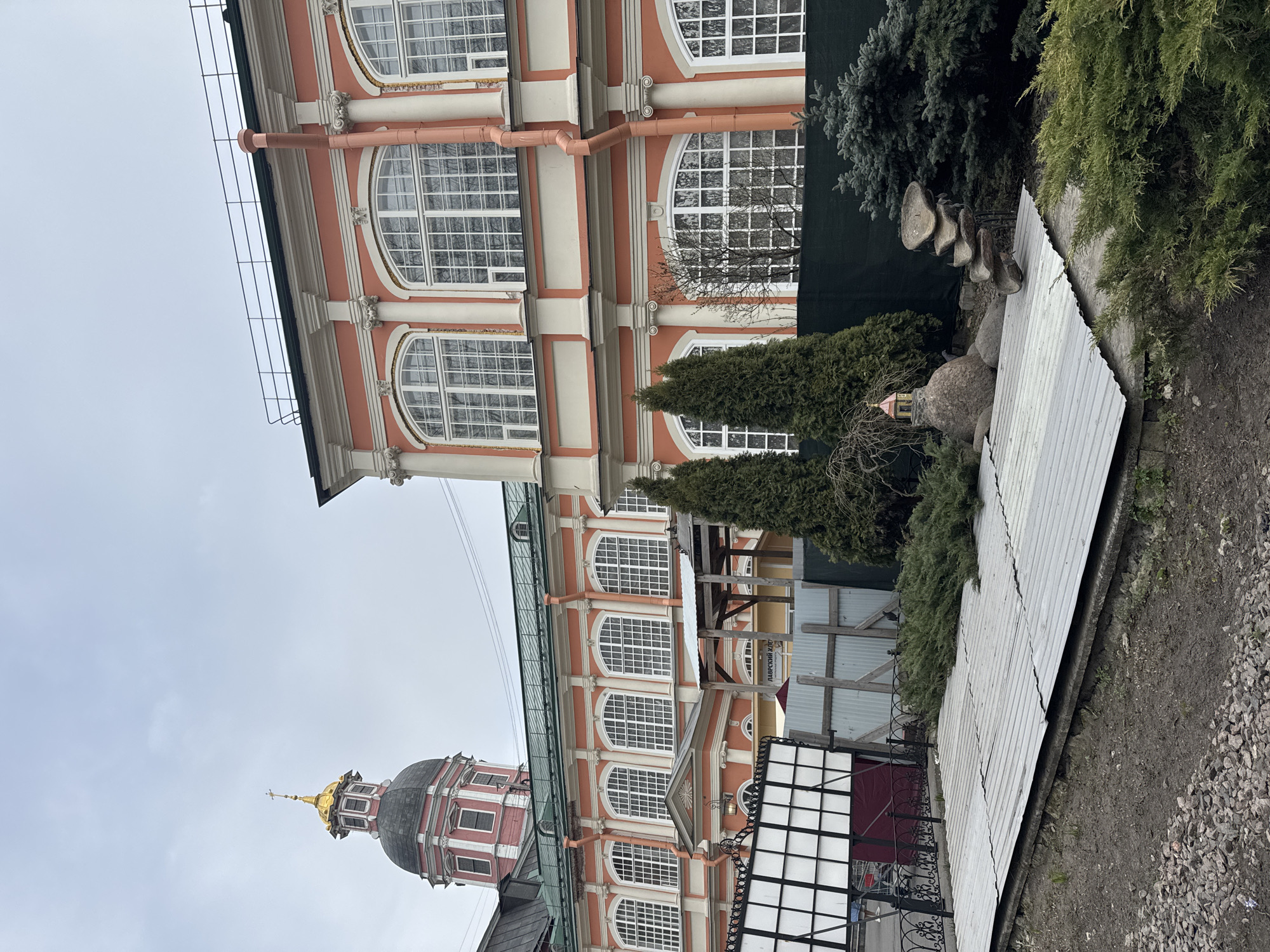 The image depicts a section of a building in St. Petersburg, Russia, characterized by its red and white facade with arched windows. A golden dome is visible in the lower left corner of the frame. The surrounding environment includes greenery and a corrugated metal barrier. The scene appears to be taken during overcast conditions.