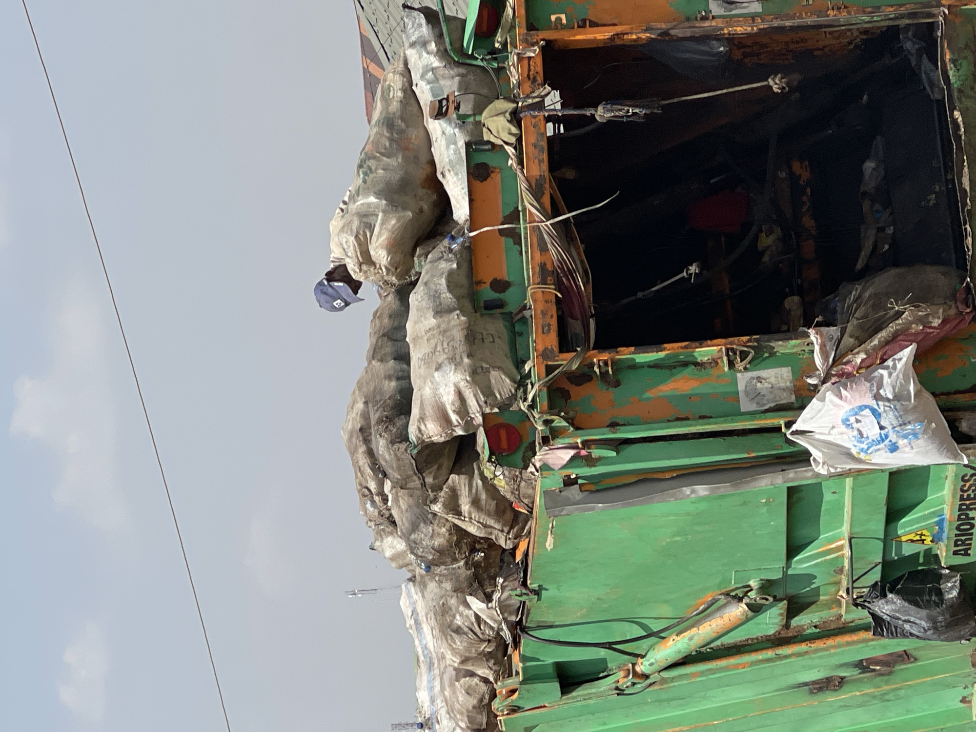 This image captures a heavily laden, weathered green and orange truck, likely involved in waste collection or material transport, under a bright, overcast sky. The truck is overflowing with numerous large, greyish-white bags, which are stacked high and appear to be tied to its side, particularly on the left. These bags likely contain collected waste or recyclable materials.
A person, wearing a blue baseball cap, is visible on top of the bags on the truck's left side, suggesting they are a worker involved in loading or securing the cargo. The truck itself shows significant wear, with rust, peeling paint, and visible hydraulic components, including a green cylinder on the lower right. The rear compartment of the truck is dark and appears to contain more assorted items, with a small red object faintly discernible within.
On the green side panel towards the right, the word "ARIOPRESS" is printed vertically in white. A smaller white bag with a faded blue logo or text is hanging near the rear right side, and a black plastic bag is also visible below it. Thin power lines or cables stretch across the hazy sky in the background. The scene depicts a moment of industrious activity related to resource management, typical of daytime operations in urban environments like Abuja, Nigeria.