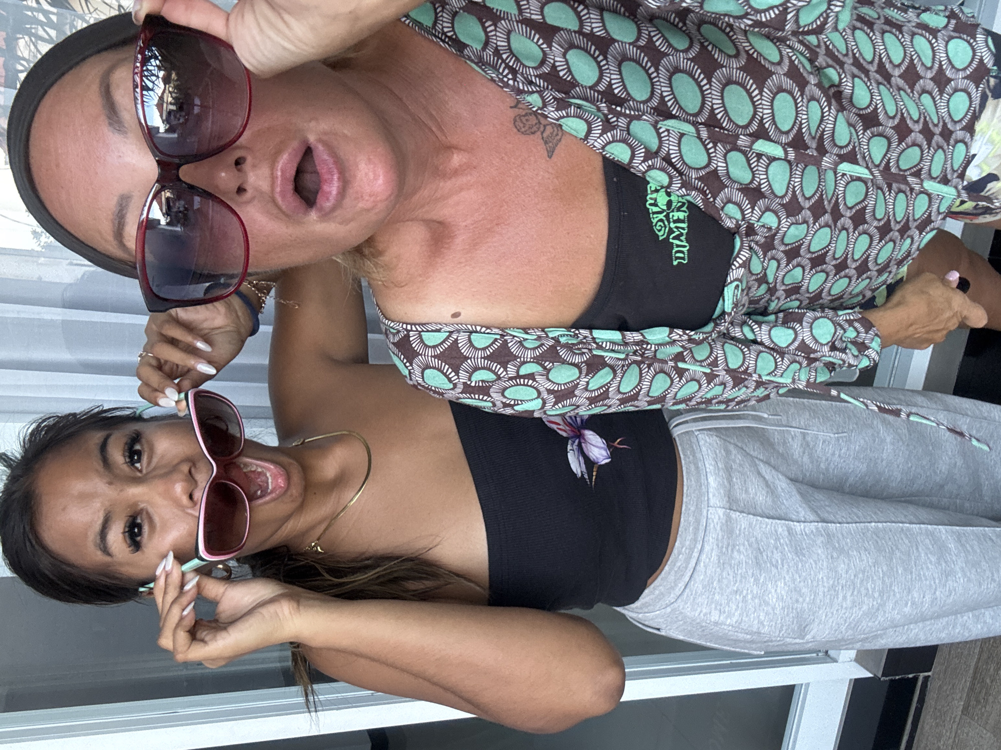 The image captures two women posing playfully, likely indoors or in a semi-enclosed space, under bright daytime lighting in Kuta, Indonesia.
The woman positioned on the left (when viewed upright) has fair skin and wears a dark headband. She is holding a pair of large, dark red-framed sunglasses slightly away from her face with her right hand, revealing her wide-open mouth in a playful expression. She is dressed in a long-sleeved top with a distinctive pattern of light green oval shapes on a dark brown background, worn over a black undergarment. On this black top, partially visible in green stylized text, are the words "OTHER DIMEN". A small, dark tattoo, possibly depicting a berry cluster, is visible on her upper chest. Her nails are manicured in a light color.
The woman on the right (when viewed upright) has darker skin and is also holding pink and light blue-framed sunglasses slightly off her face with both hands, mirroring the first woman's wide-open mouth and cheerful expression. She wears a simple gold chain necklace, a black strapless crop top, and light grey sweatpants. A stylized purple and white floral or plant emblem is visible on the front of her black top. Her nails feature white tips.
Both women appear joyful and energetic, engaging directly with the camera. The background is minimal, showing bright reflections, possibly from windows or a light-colored wall, and some darker floor or wall sections. The overall scene suggests a casual and fun moment captured between the two individuals.
