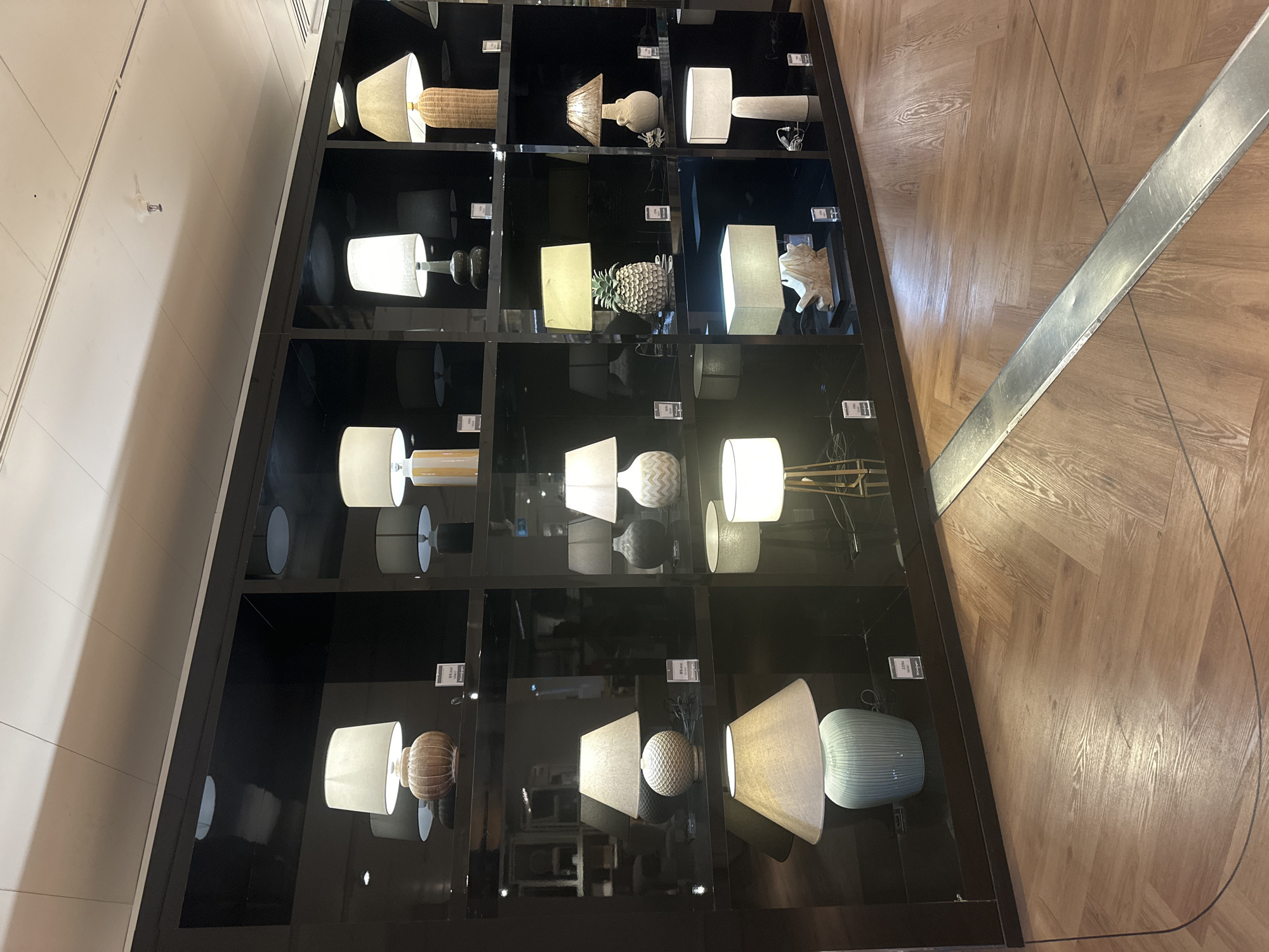 In a retail store environment, a wall display showcases an assortment of table lamps. The lamps are arranged on dark, reflective shelves against a black background, suggesting a modern and minimalist aesthetic. The display features a variety of lamp designs, including those with textured bases, cylindrical shades, and decorative elements such as a pineapple-shaped lamp. Some lamps are illuminated, casting a warm glow on the surrounding surfaces. The flooring in the foreground is made of light-colored wooden planks laid in a herringbone pattern. The overall scene depicts a well-lit retail space designed to highlight home decor items. The location is in Barcelona, Spain, as indicated by the price tags on some of the lamps, which are in Euros.